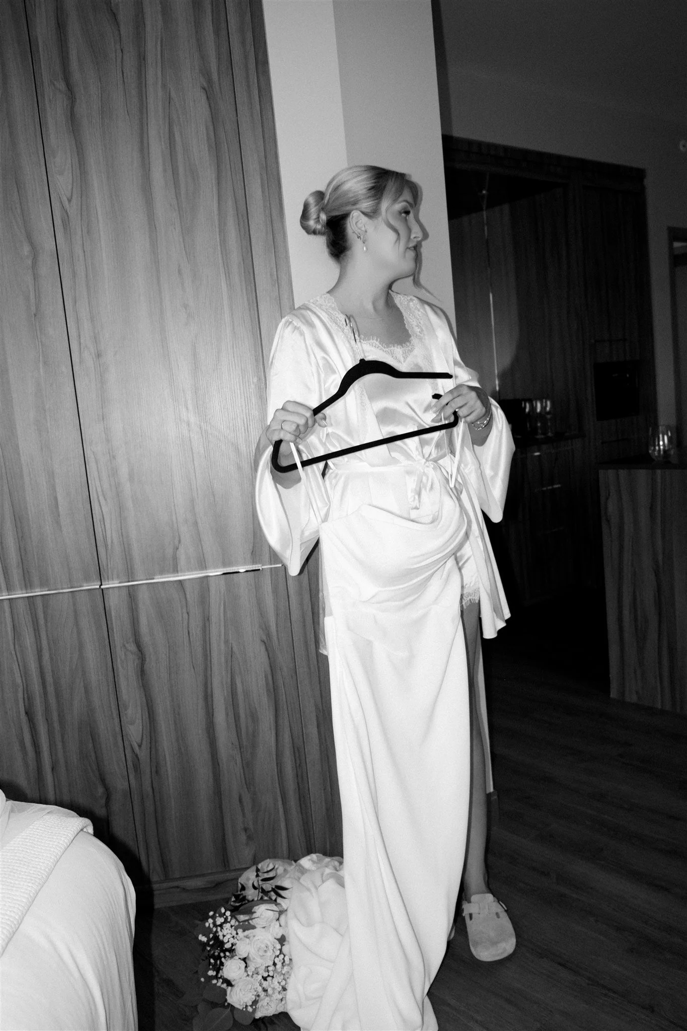 Une femme habillée d'une robe de mariée blanche, se préparant dans une pièce intérieure avec un décor en bois, tenant un cintre dans une main, une bouquet de fleurs blanc posé au sol.