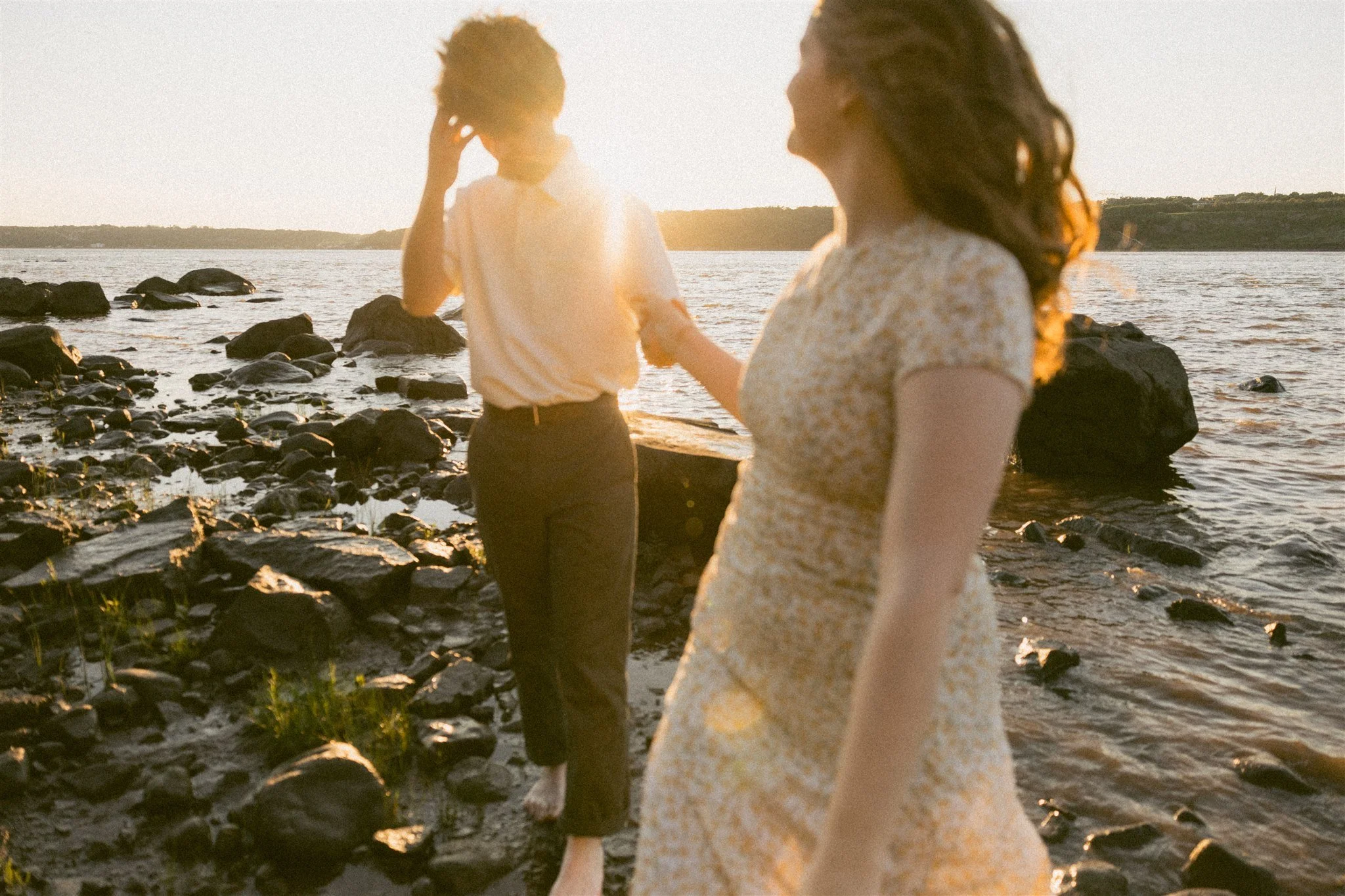 Deux femmes sur une plage rocheuse au coucher du soleil, l'une regarde l'eau tandis que l'autre regarde la première.
