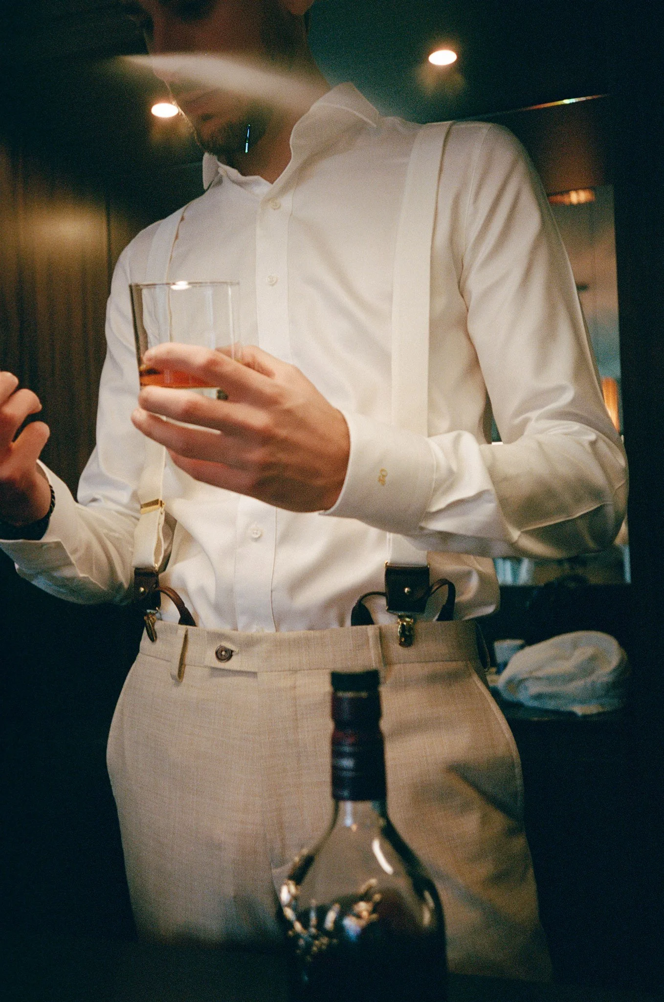Homme portant une chemise blanche et des bretelles, tenant un verre de whisky, dans une ambiance sombre.