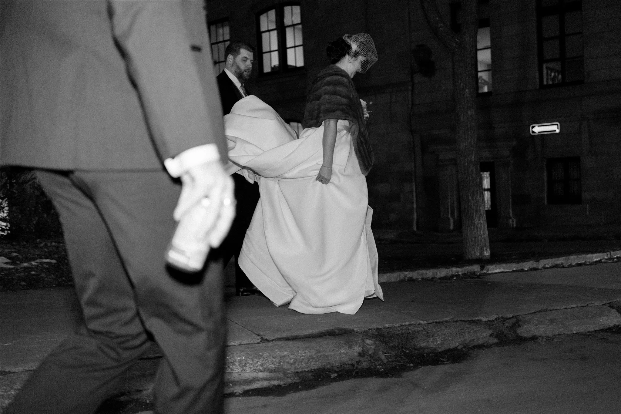 Une scène de mariage en noir et blanc avec une femme en robe de mariée et un homme, probablement le marié, à l'arrière-plan. La femme porte un voile et une cape, marchant sur le trottoir dans une rue urbaine.