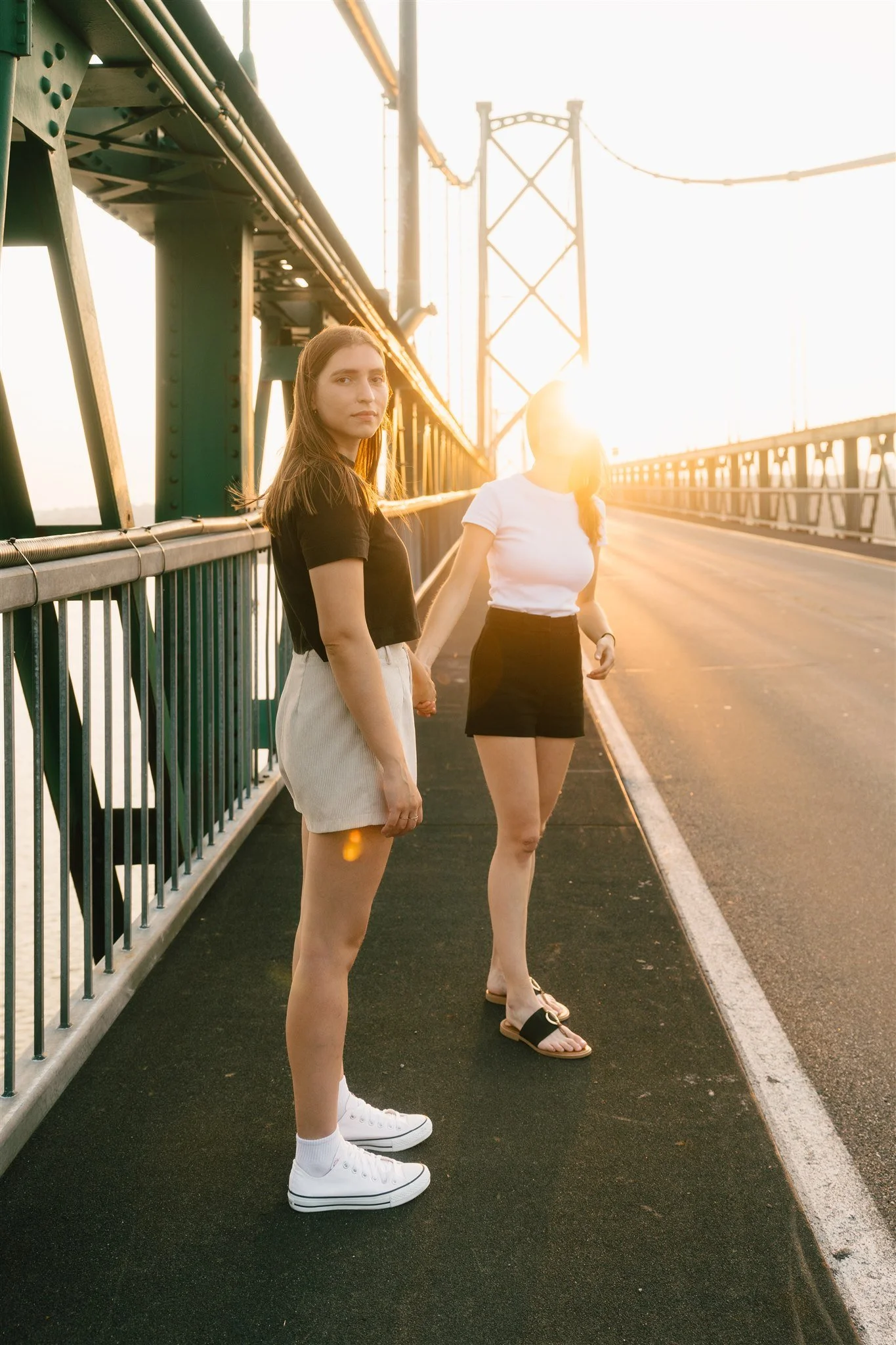 Deux femmes debout sur un pont au coucher du soleil, tenant la main, l'une portant une robe noire et une jupe blanche, l'autre portant un t-shirt blanc et une jupe noire, avec des chaussures décontractées.