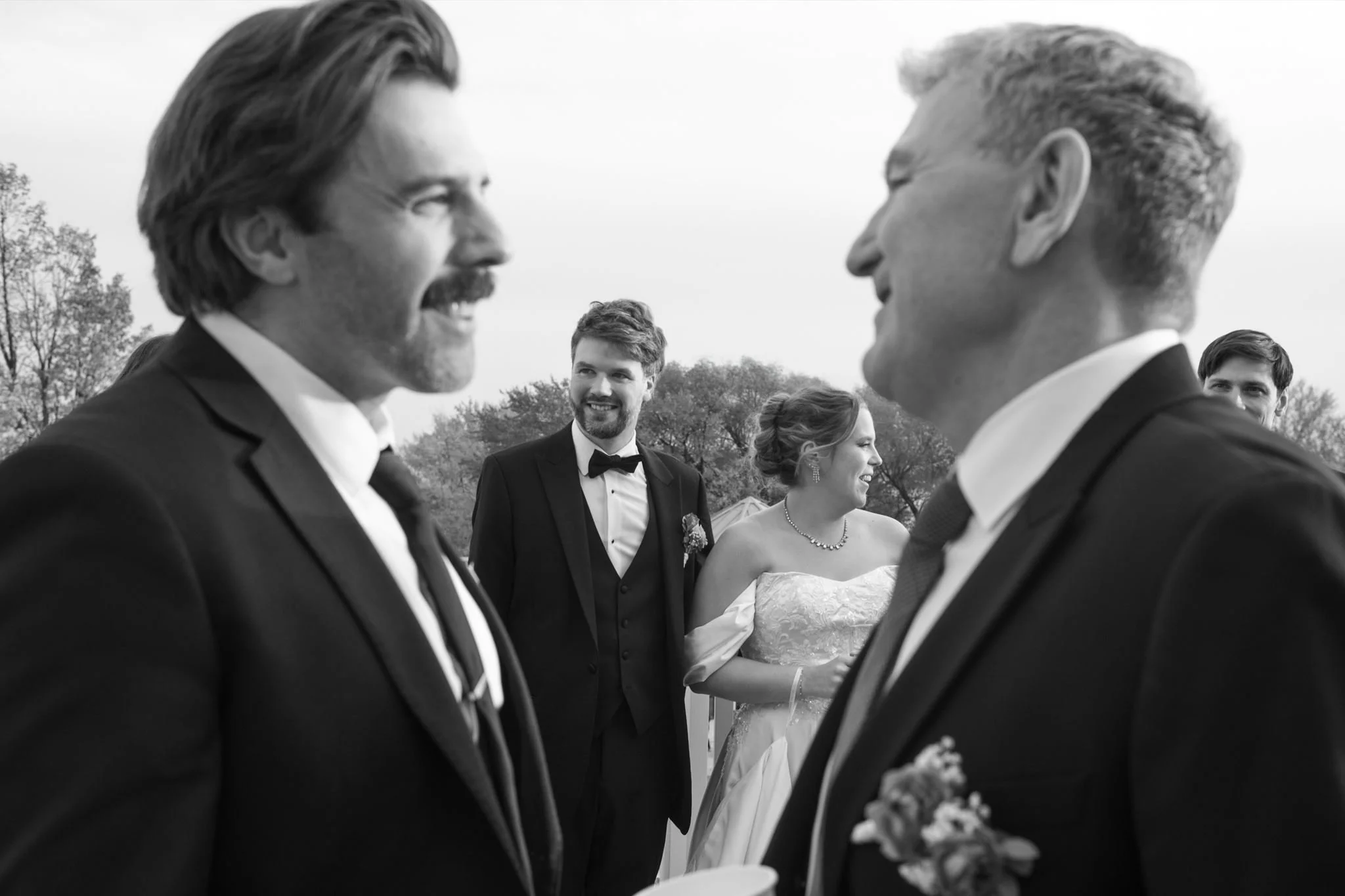 Groupe de personnes lors d'un mariage en extérieur, en noir et blanc, souriants et habillés en tenue formelle.