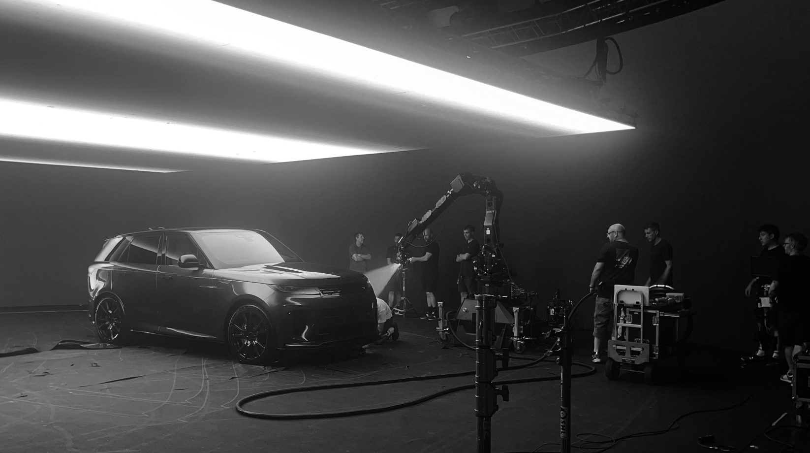 Black and white photo of a modern SUV being filmed with multiple crew members and filming equipment in a dark studio