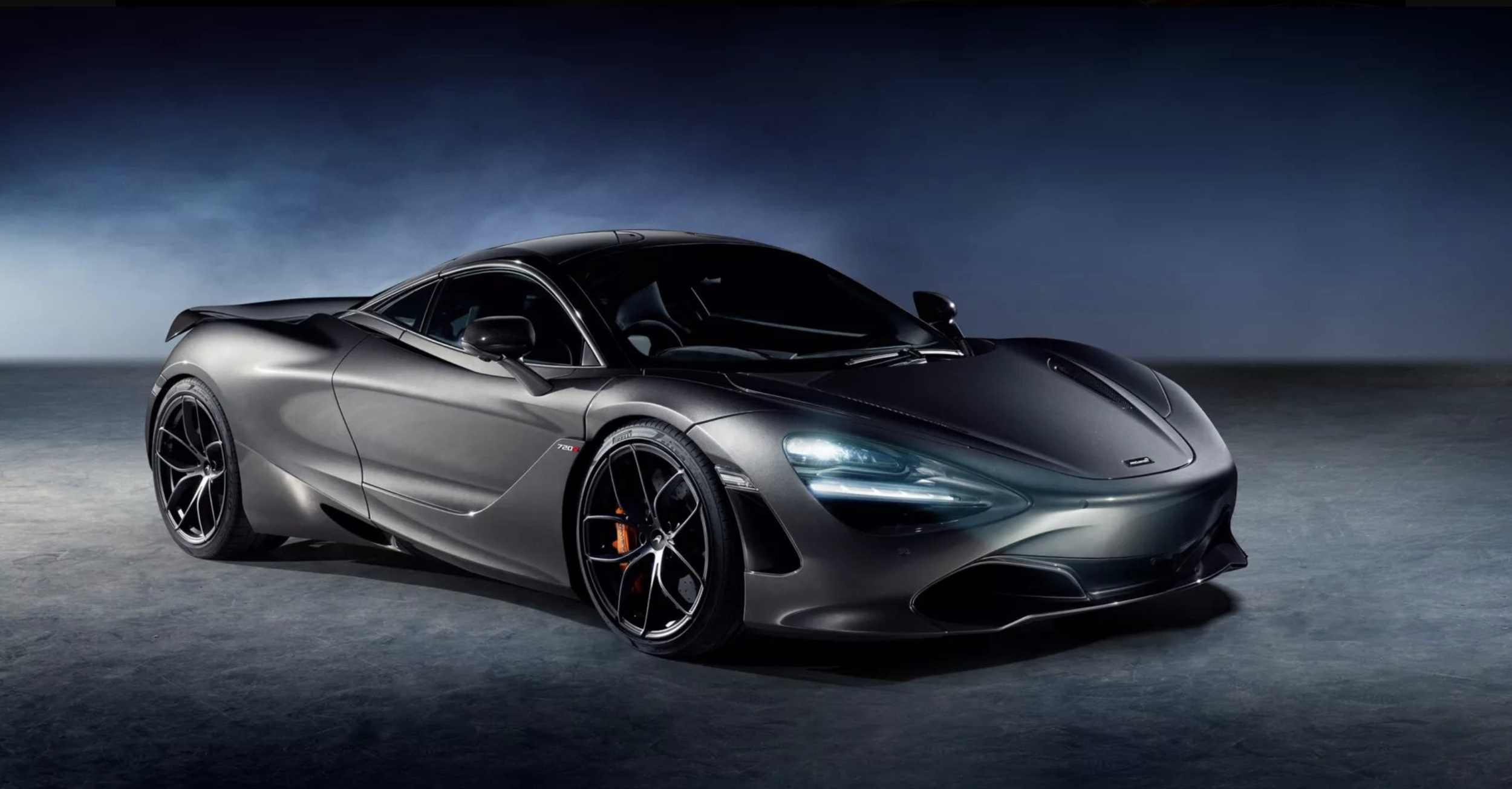 A sleek, black sports car parked on a dark surface against a moody, cloudy sky background at night.
