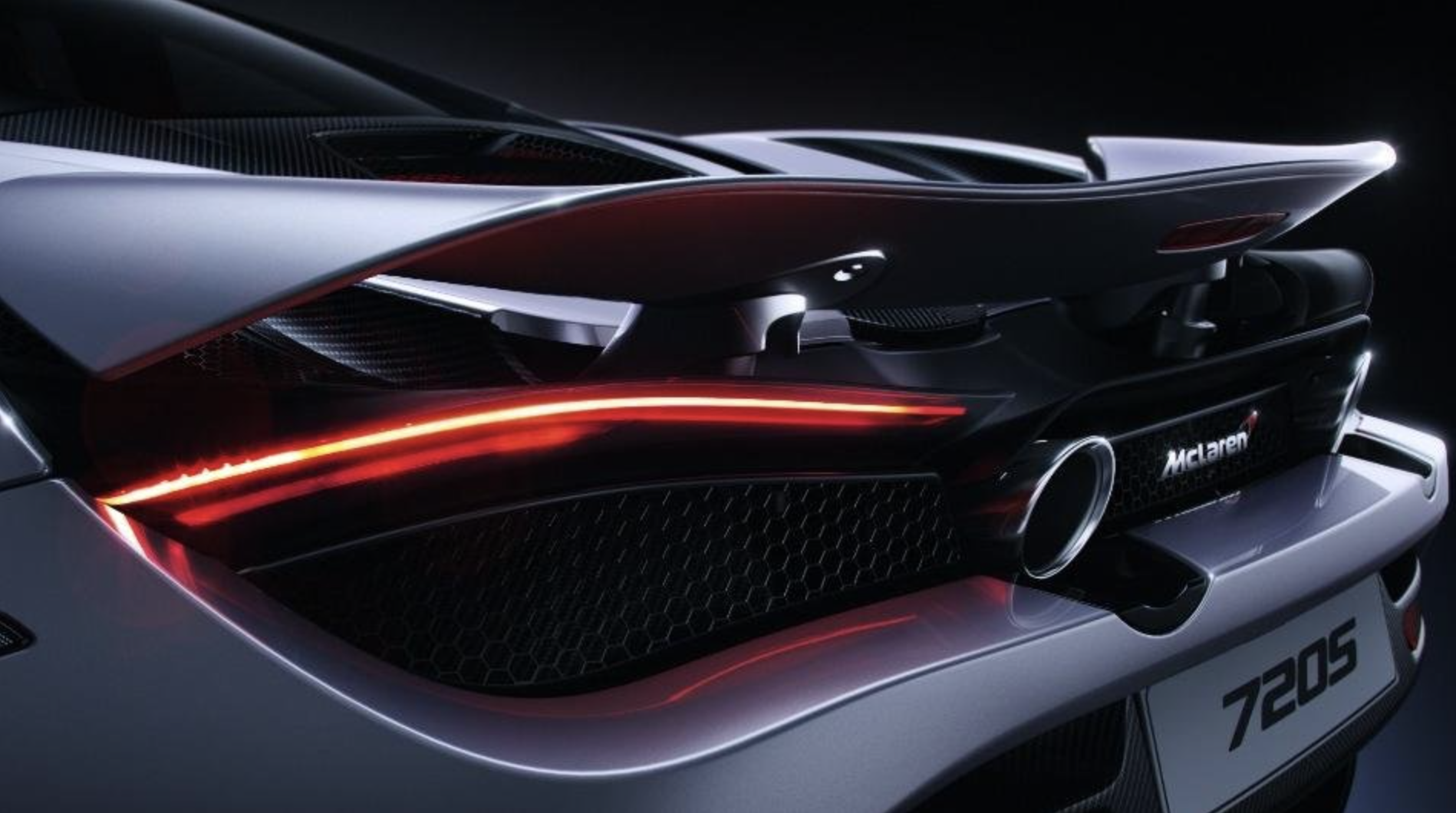 Close-up of the rear of a silver McLaren 720S sports car, featuring aerodynamic design, a distinctive taillight with a red light strip, a black mesh vent, a large exhaust pipe, and McLaren branding.