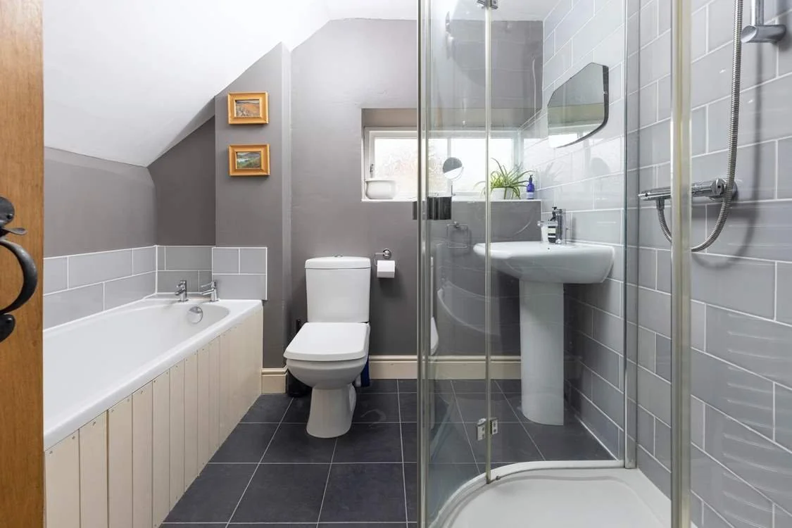 Small bathroom with a bathtub, toilet, and shower stall with glass door, gray tiled floor, and gray walls with small framed pictures.