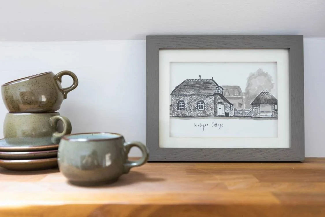 A wooden surface holding a stack of four ceramic cups and saucers, and a framed black-and-white drawing of a cottage labeled "Halagon Cottage."