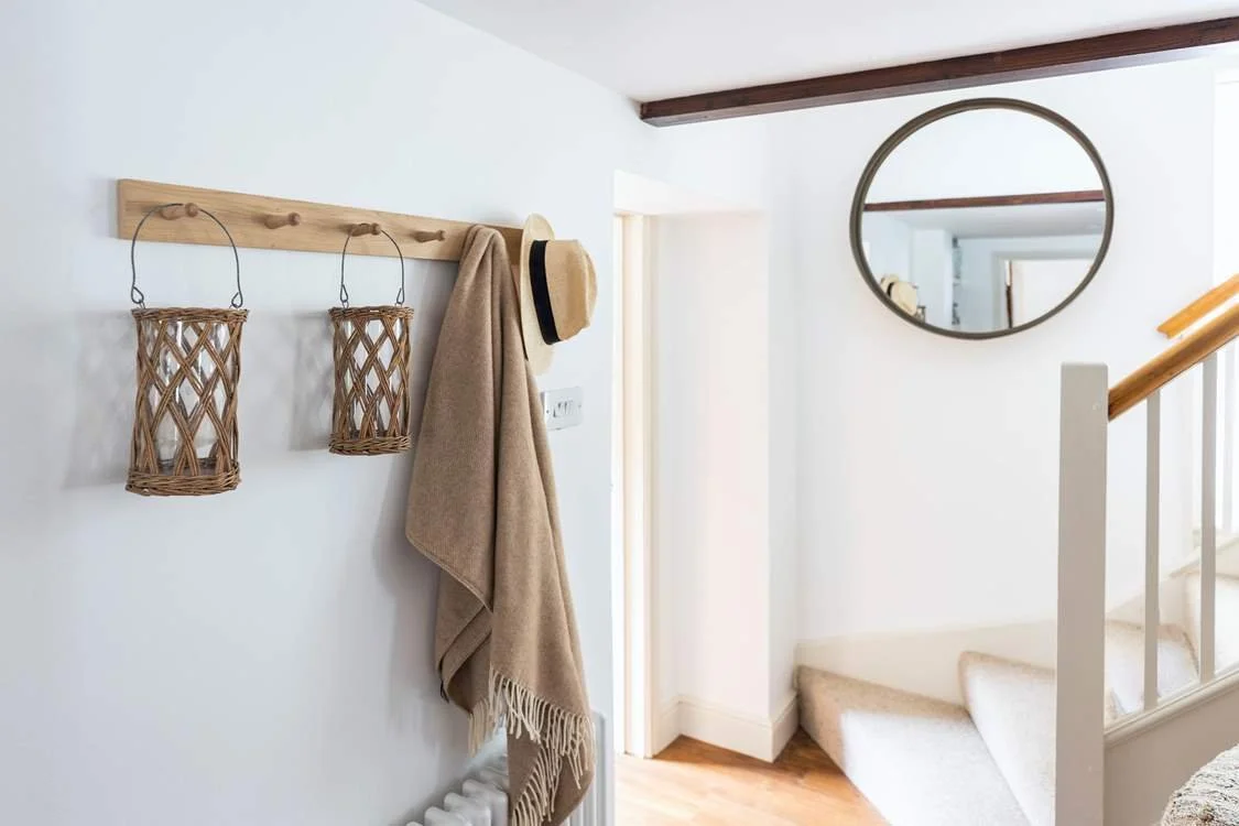 Hallway with white walls, wooden coat rack with woven lanterns, beige coat, beige hat, circular mirror, staircase with white railing and beige carpet.