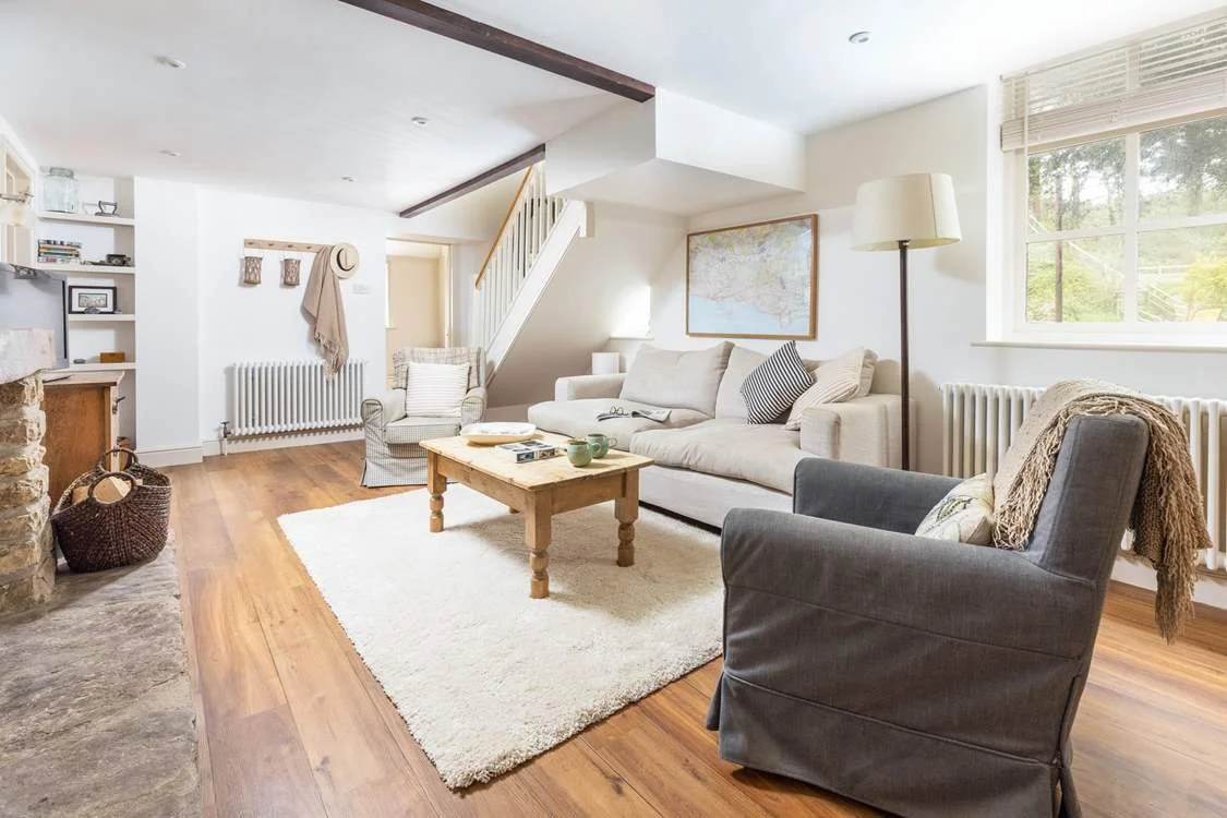 Bright living room with white walls, wooden floors, a white sofa, armchair, coffee table, and large window with blinds.