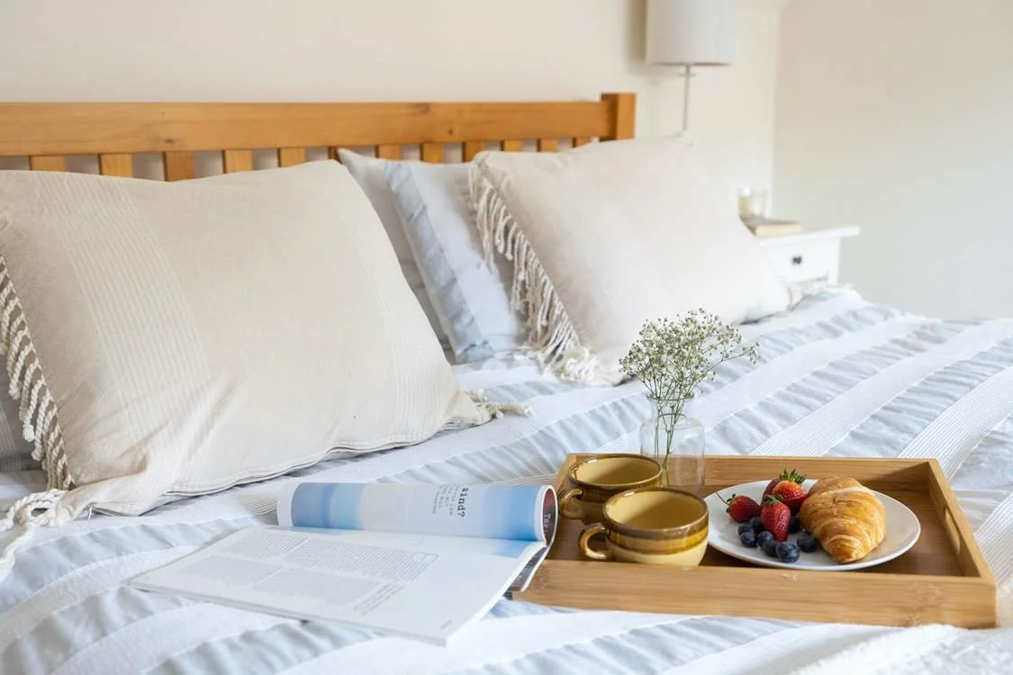 A neatly made bed with cream-colored pillows, a wooden headboard, a bedside lamp, and a tray with a vase of flowers, two cups, an open magazine, and a plate of strawberries, blueberries, and a croissant.