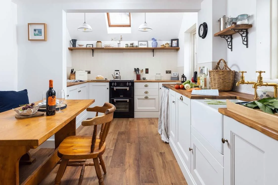 A bright, cozy kitchen with white cabinets, wooden countertops, and a wooden dining table with chairs. There are hanging lights, a skylight, and open shelves with jars and decorations.