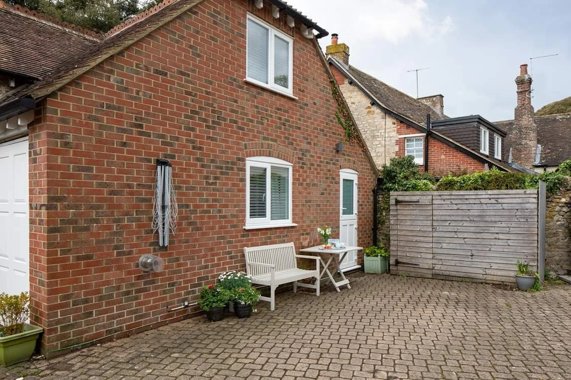 Halcyon Cottage is a small brick house with a concrete patio, white bench, table with flowers, and a wooden privacy screen, in a suburban neighborhood.