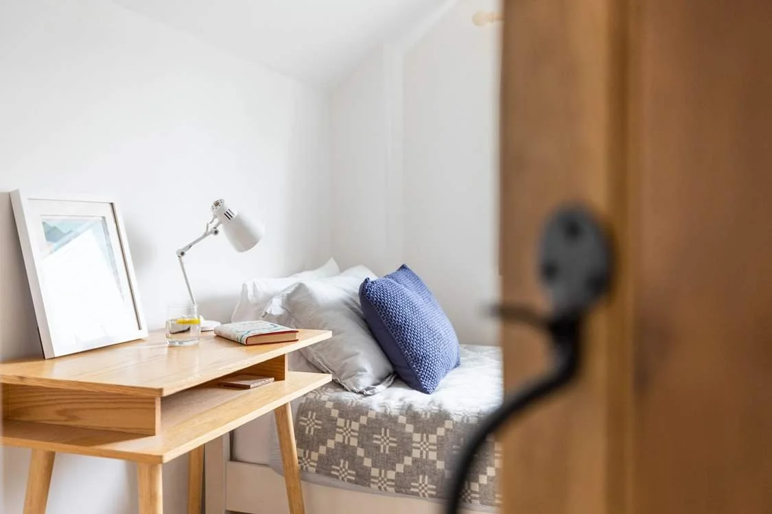 Photo of a bedroom with a bed, blue pillow, wooden side table, framed picture, and a desk lamp, seen through an open door.