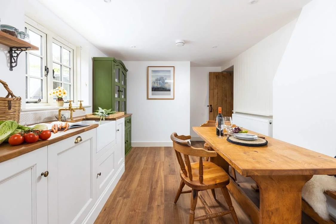 A bright kitchen with white cabinets, wooden countertops, and a wooden dining table with chairs. Green cabinet, window with flowers, and wooden floor.