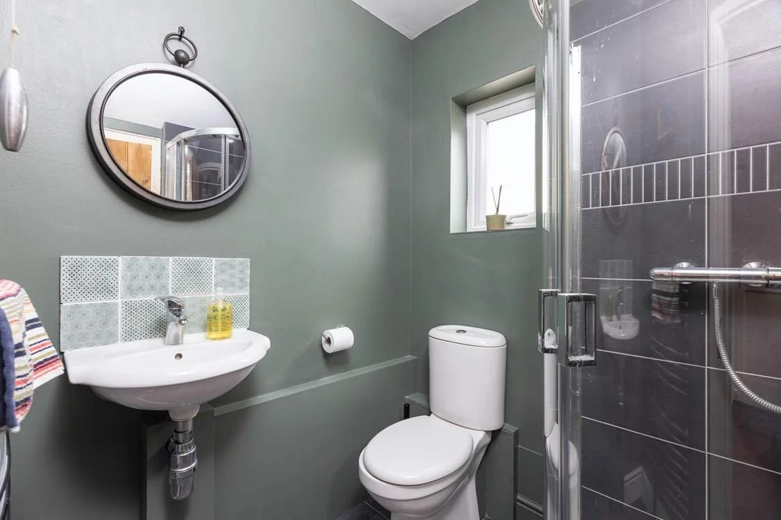 Small bathroom with a wall-mounted sink, a toilet, a window with a small plant, a round mirror, and a shower with black tiled walls.