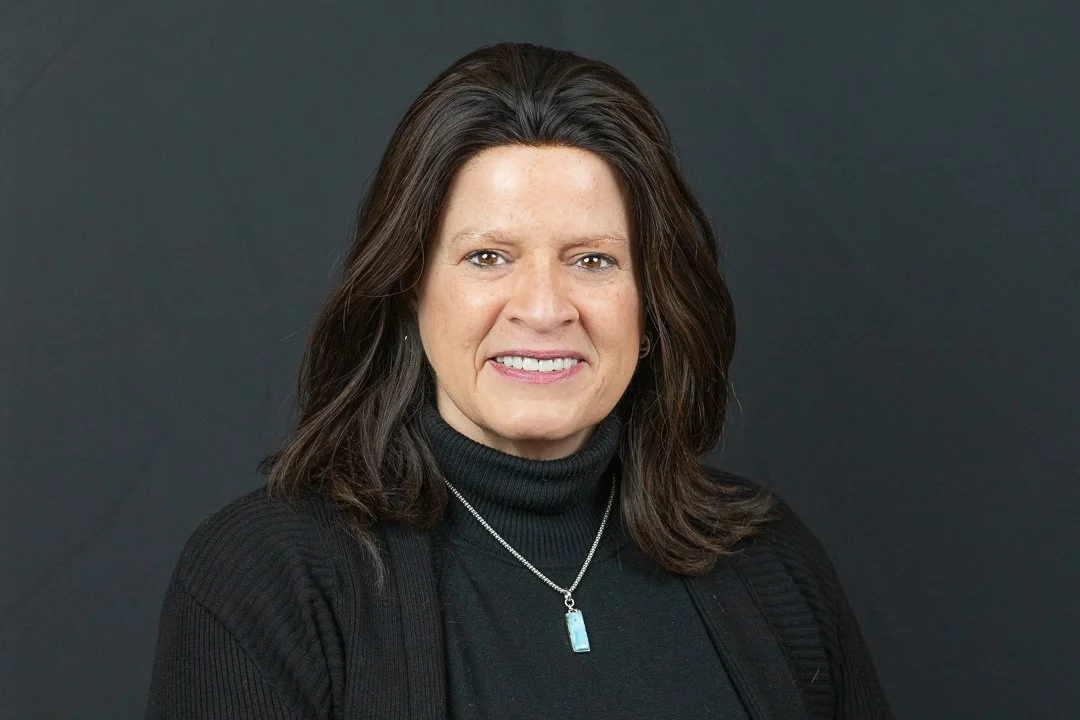 A woman with shoulder-length dark brown hair, wearing a black turtleneck, a black cardigan, and a silver necklace with a blue pendant, smiling against a plain dark background.
