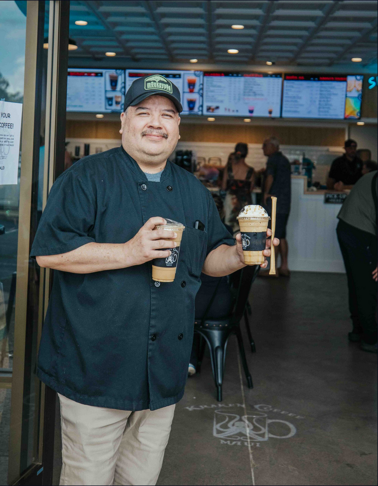 A man in a black chef's coat, beige pants, and a baseball cap standing outside a cafe, holding two cold coffee drinks and smiling.