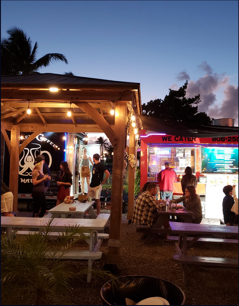 An outdoor dining area at a sunset with picnic tables, a food stand, and a coffee truck, illuminated by string and neon lights, with people eating and socializing.