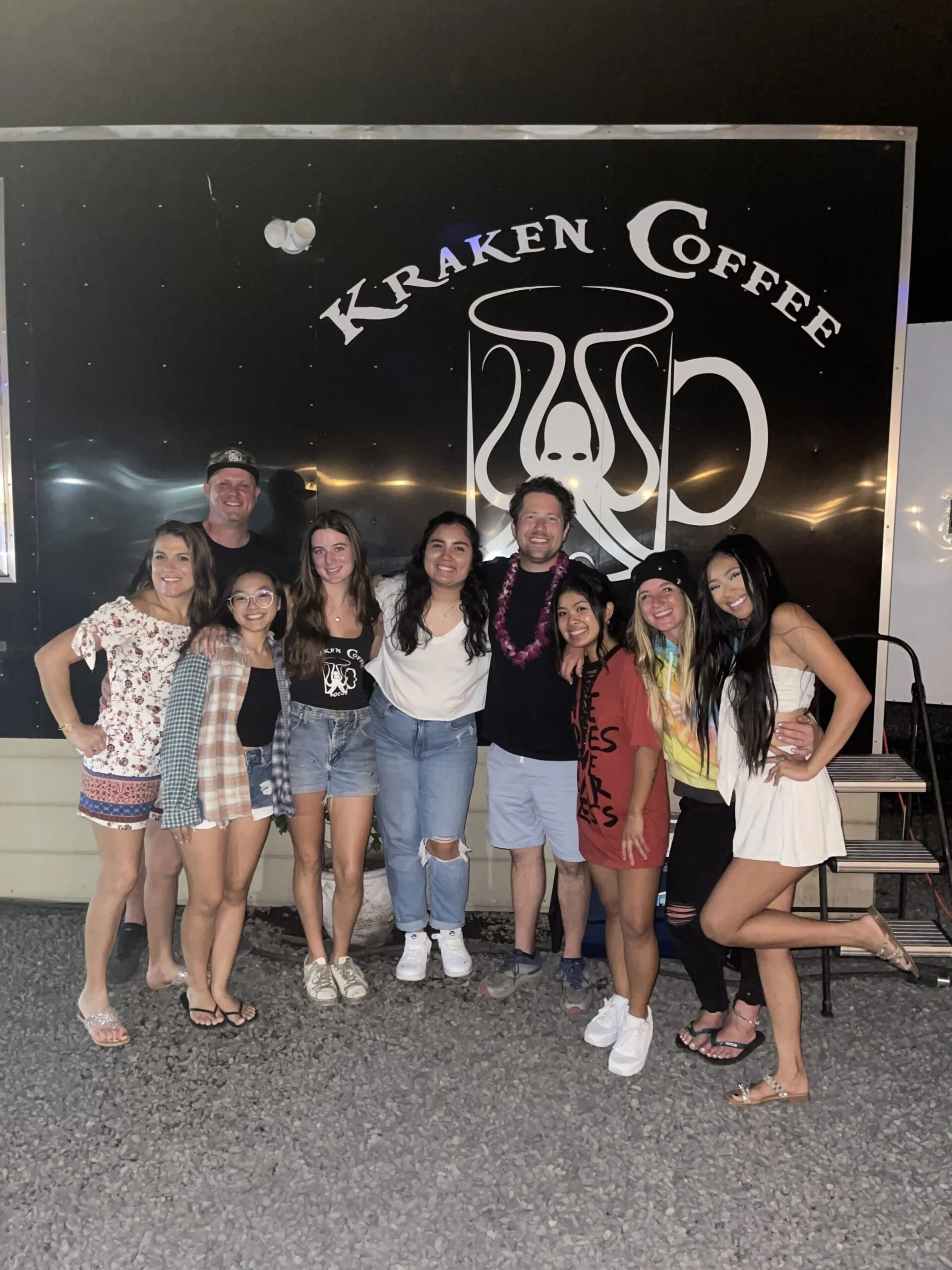 Group of nine people posing in front of Kraken Coffee sign at night, outdoors.