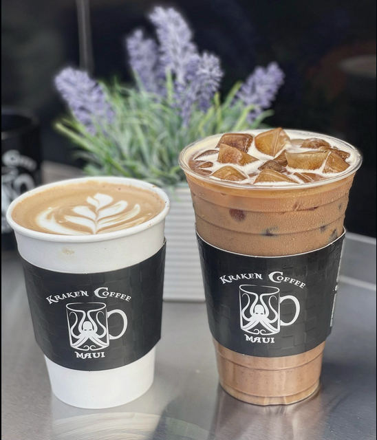 Two iced coffee drinks in black and white paper cups with Kraken Coffee Maui logos, one topped with latte art, the other with ice cubes, with purple flowers in a white container in the background.
