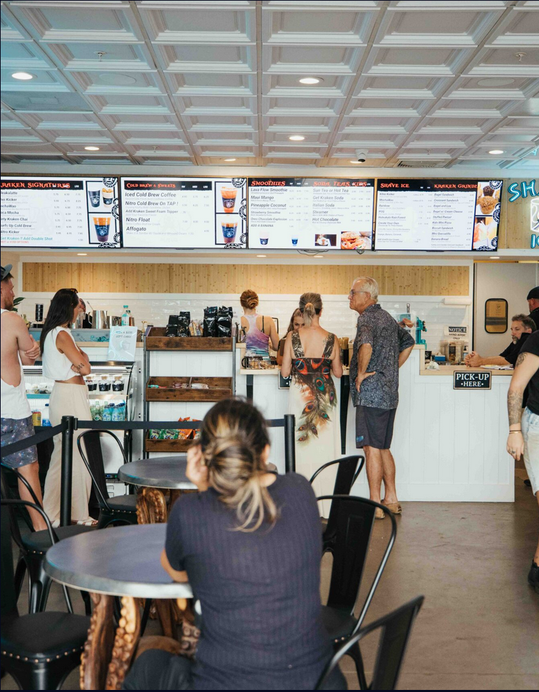 People ordering and waiting at a counter in a cafe or ice cream shop, with menu boards displayed above.