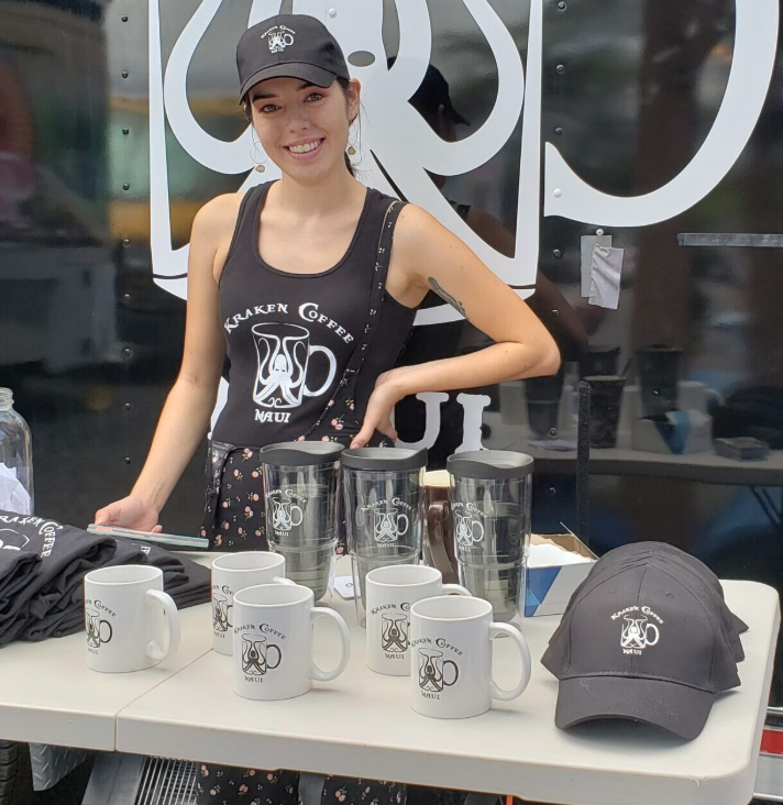 A smiling woman wearing a black tank top, black cap, and earrings standing behind a table with coffee mugs, tumblers, and black hats, at a coffee promotional booth for Kraken Coffee Maui.