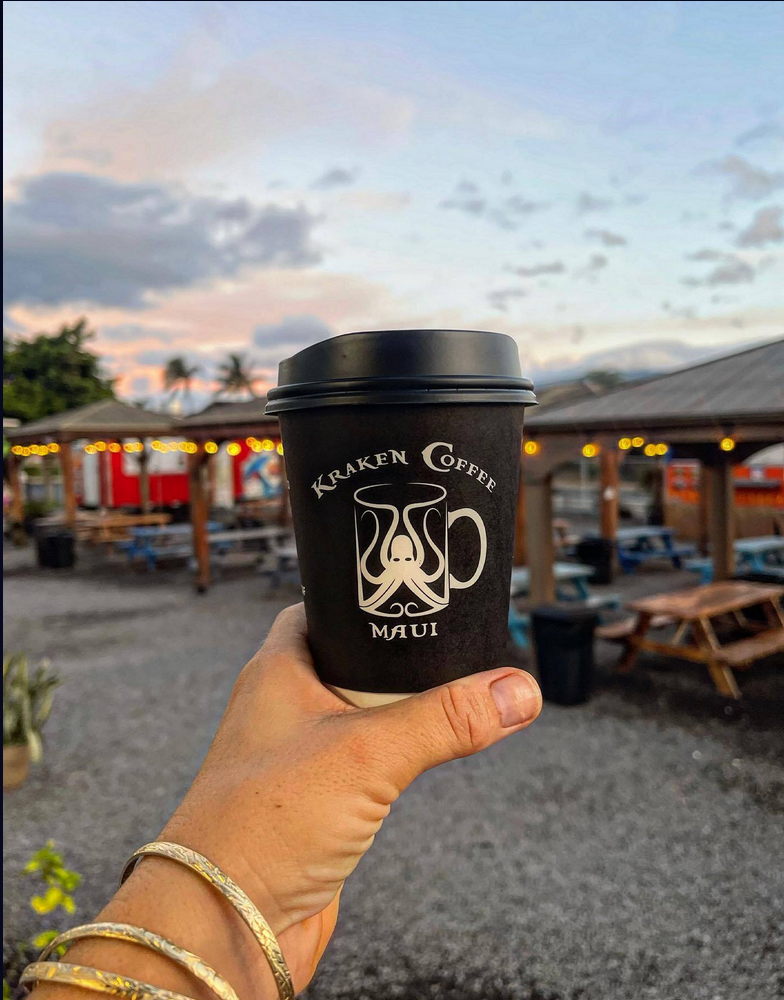 Hand holding a black paper coffee cup with a logo that says 'Kraken Coffee Maui,' outside seating area with picnic tables and a small structure, sunset sky with clouds in the background.