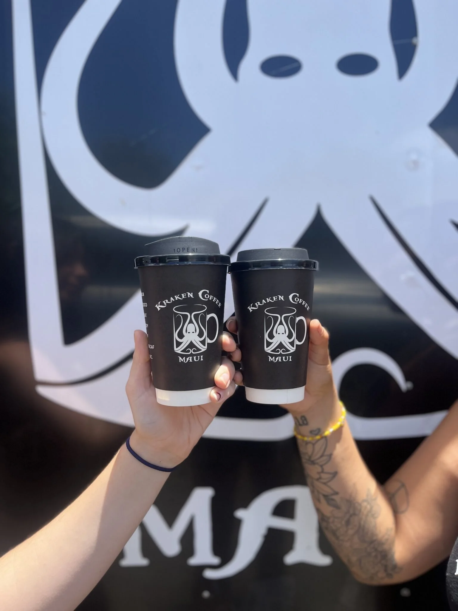 Two hands holding black disposable coffee cups with white logo from Kraken Coffee in Maui, Hawaii, in front of a black and white Kraken Coffee sign.