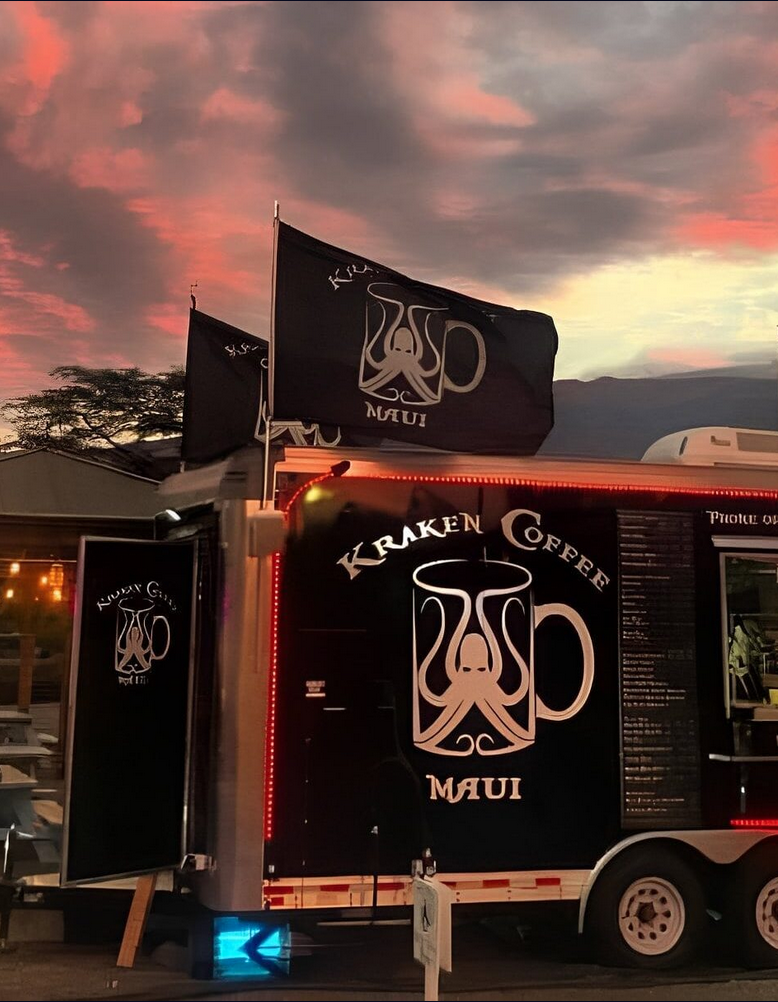 A food truck with the logo of Kraken Coffee Maui, featuring a black background, a stylized octopus coffee mug, and illuminated by red and blue lights against a sunset sky.