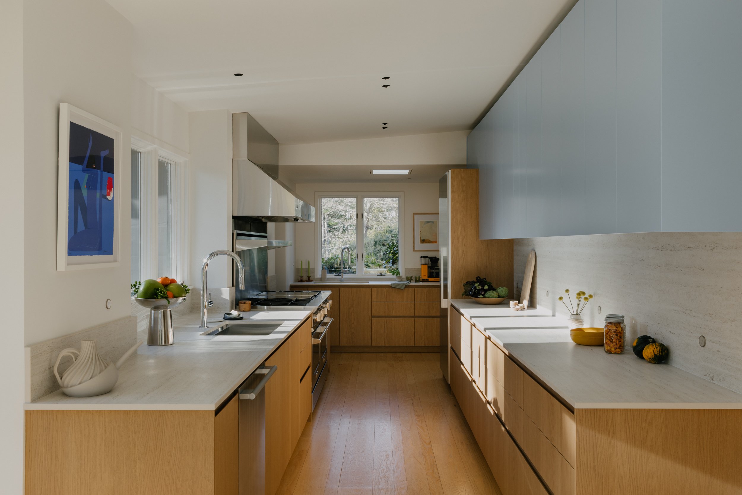 Modern kitchen with wooden cabinets, white countertops, and stainless steel appliances. Natural light from windows illuminates the space, with decorative items and vegetables on the counter.