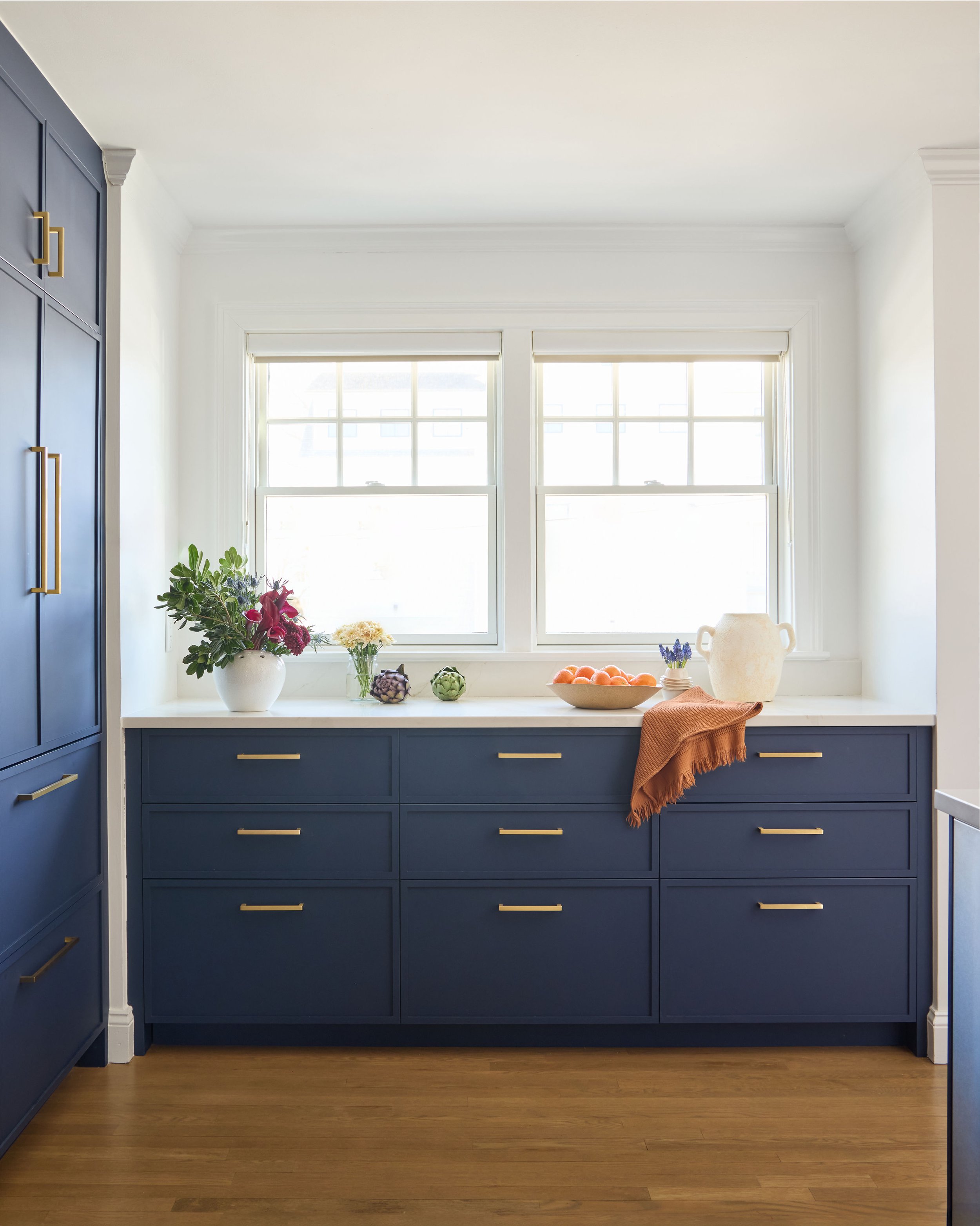 A kitchen window with blue cabinets below, decorated with flowers, fruits, and a cloth.