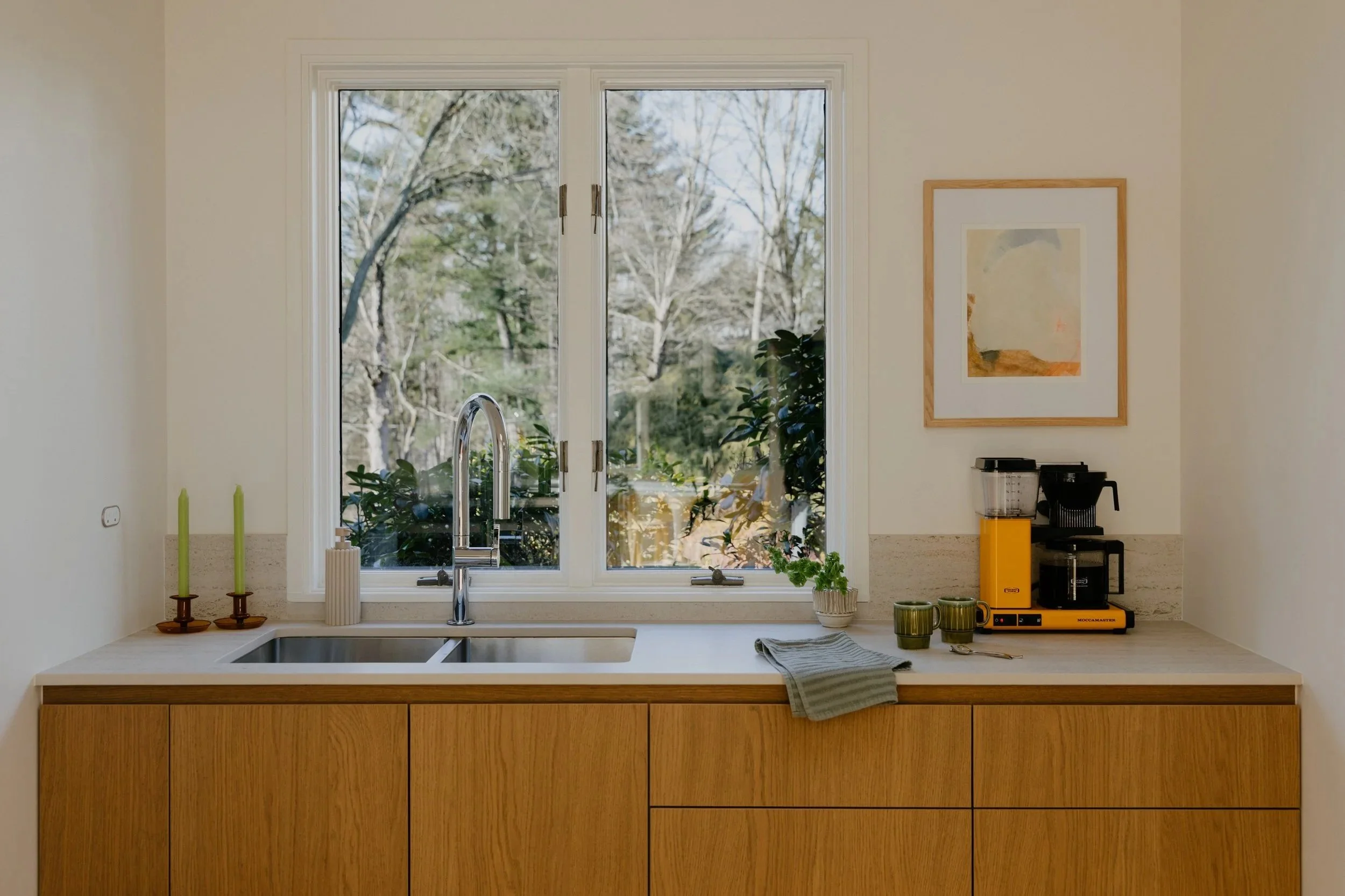 Kitchen with a large window above a wooden countertop, sink, and modern faucet. On the countertop, there are two green candles, a potted plant, a yellow coffee machine, a coffee maker, and two green mugs.