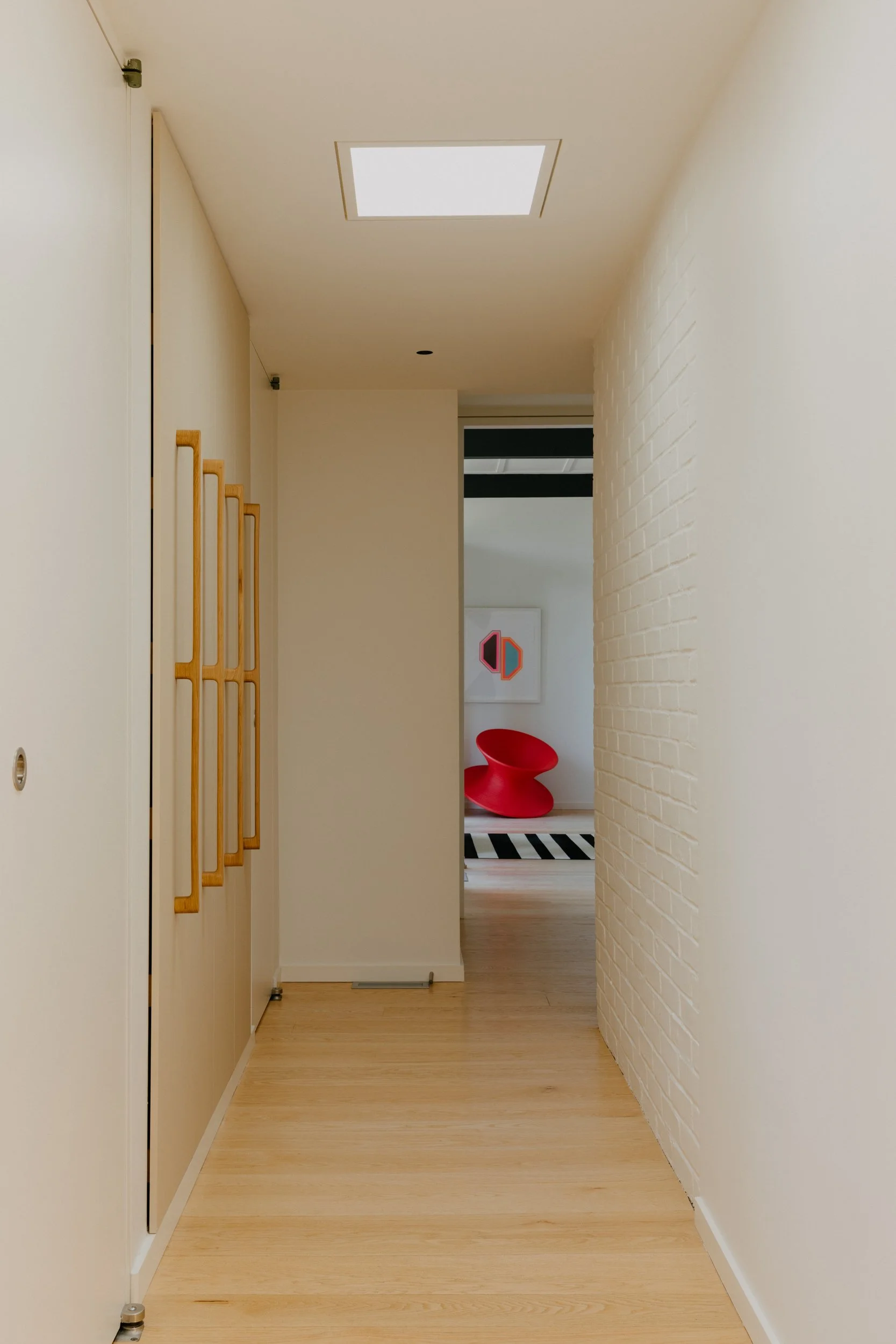 A hallway with white walls, light wood flooring, a ceiling light, and a white brick wall on the right. There are wooden hooks on the left wall, leading to a room with modern furniture and artwork, including a red chair and a black and white striped r