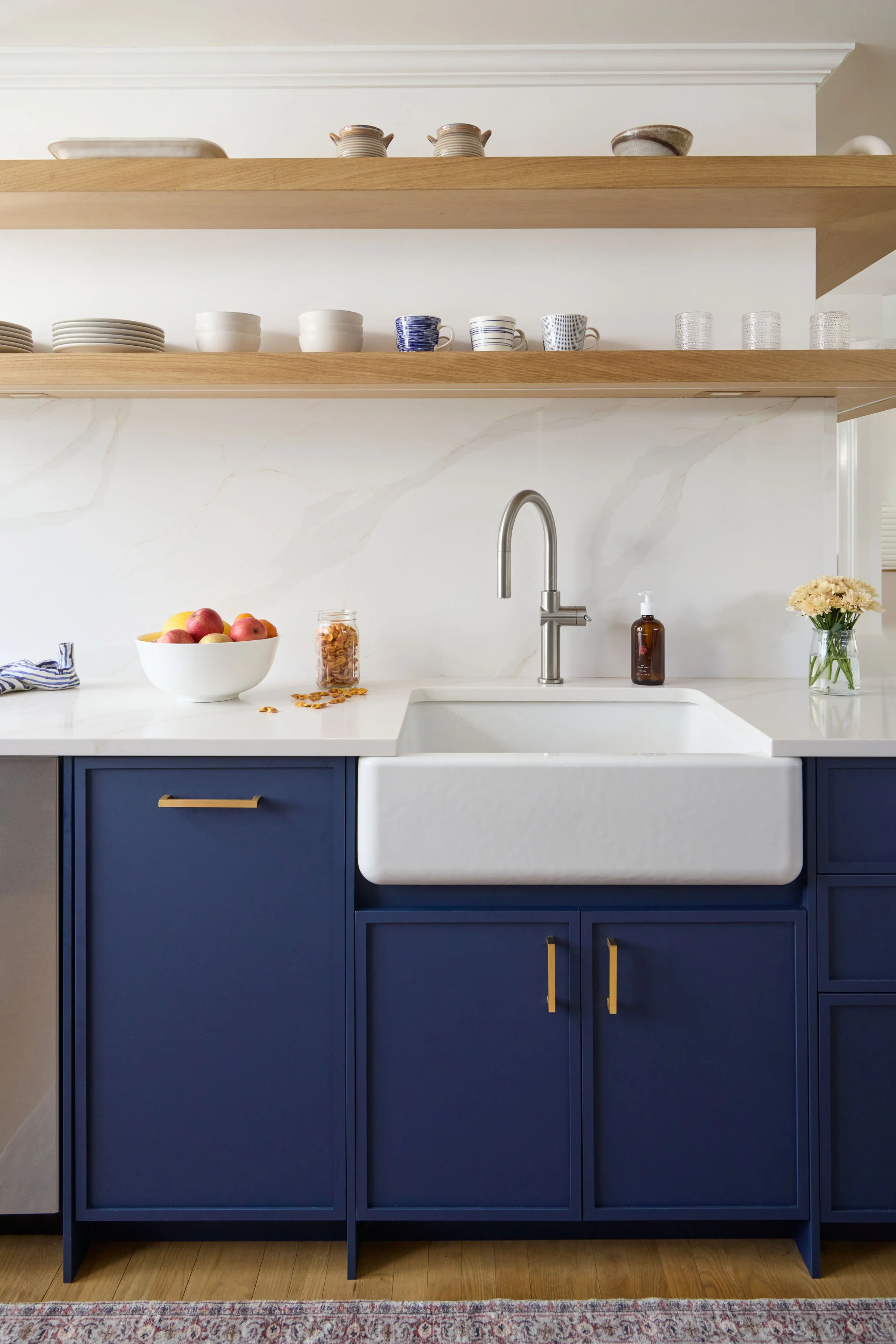 A modern kitchen with a white marble countertop, a white farmhouse sink, a stainless steel faucet, blue cabinet doors with gold handles, open wooden shelves with dishes and cups, a bowl of apples, a jar of granola, a soap dispenser, and a small vase 