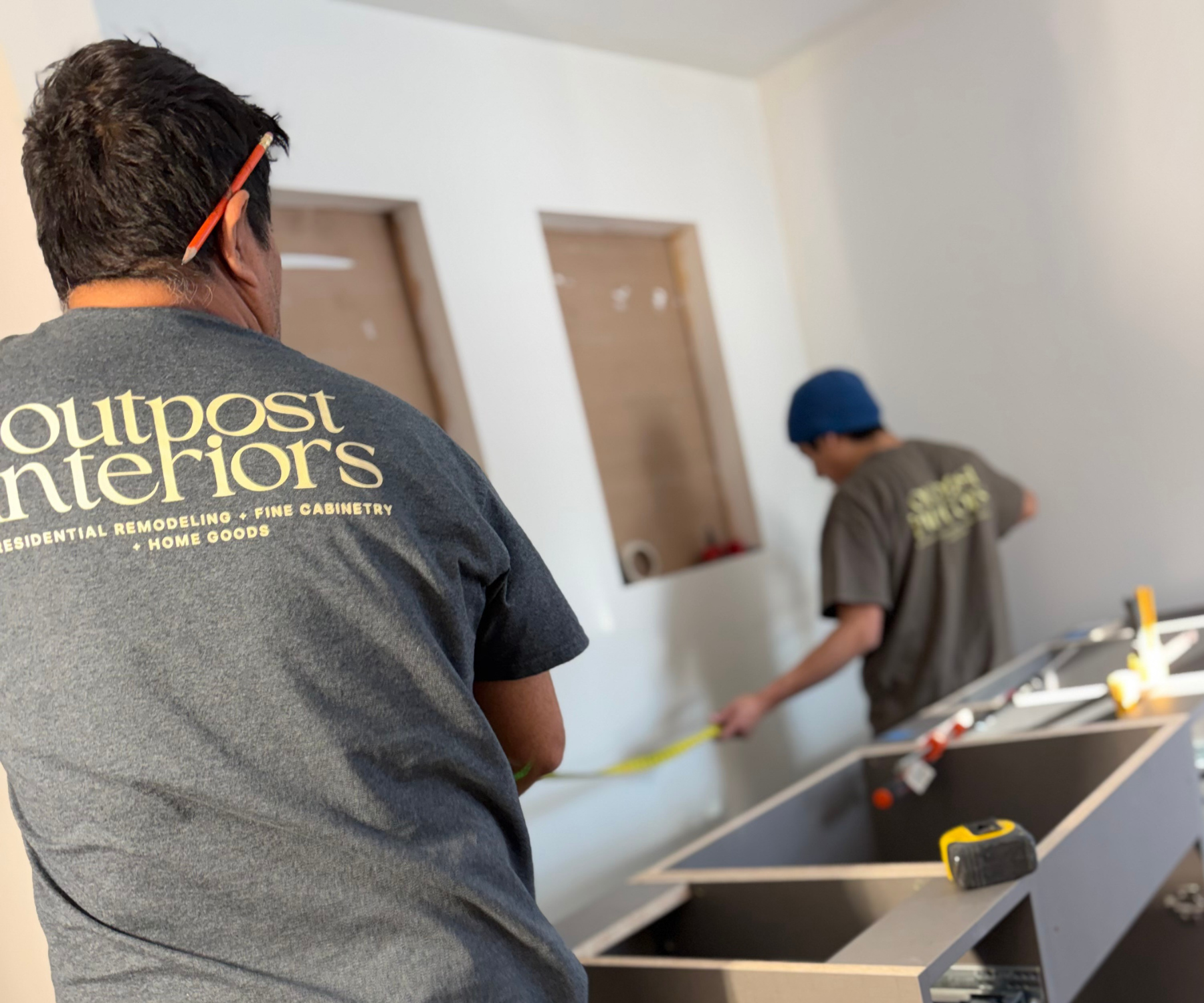 Two workers in a home renovation project measuring a wall with a tape measure. One worker is in the foreground, wearing a dark t-shirt with 'Outpost Interiors' in yellow letters, and the other worker in the background wearing a blue head covering.