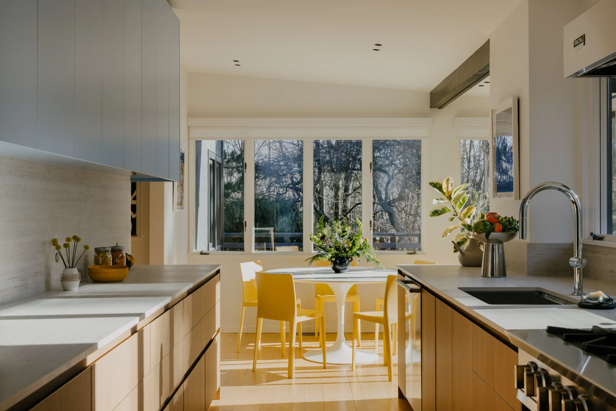 Modern kitchen with wooden cabinets, a white countertop, a gray backsplash, a sink, and a stainless steel stove. A window shows trees outside, and a small dining area with a round white table, yellow chairs, and a flower arrangement in a black vase.