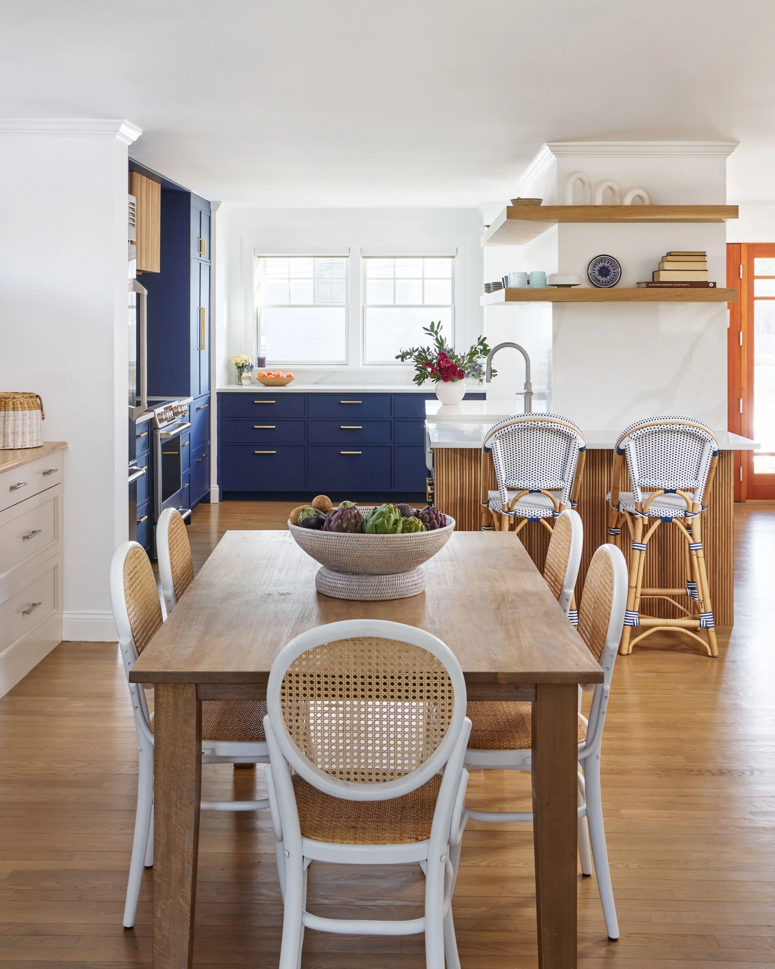 A bright kitchen with a wooden dining table and six white chairs with wicker seats. The kitchen has blue lower cabinets, white walls, and a white marble island with a faucet. There are open wooden shelves with decorative items, and a bowl of fresh ve