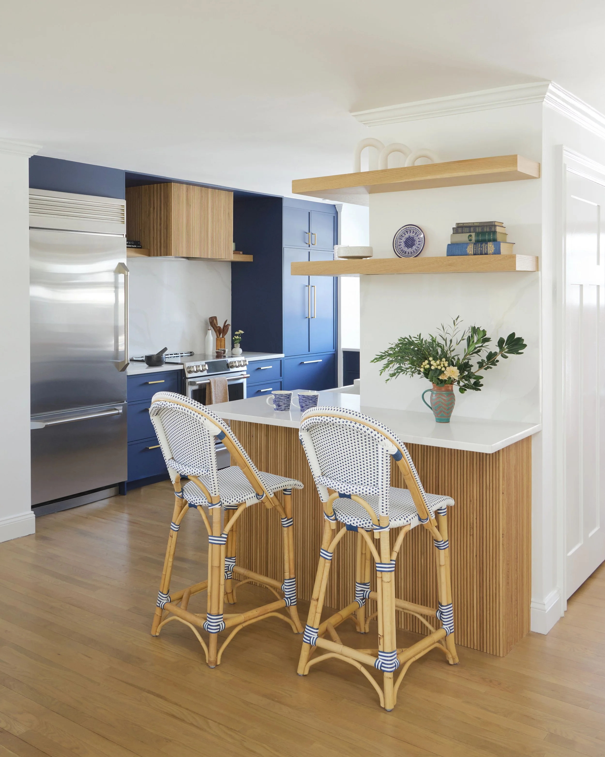 Modern kitchen with blue cabinets, wooden accents, a white countertop bar with two wicker chairs, and a vase with green foliage on the counter.