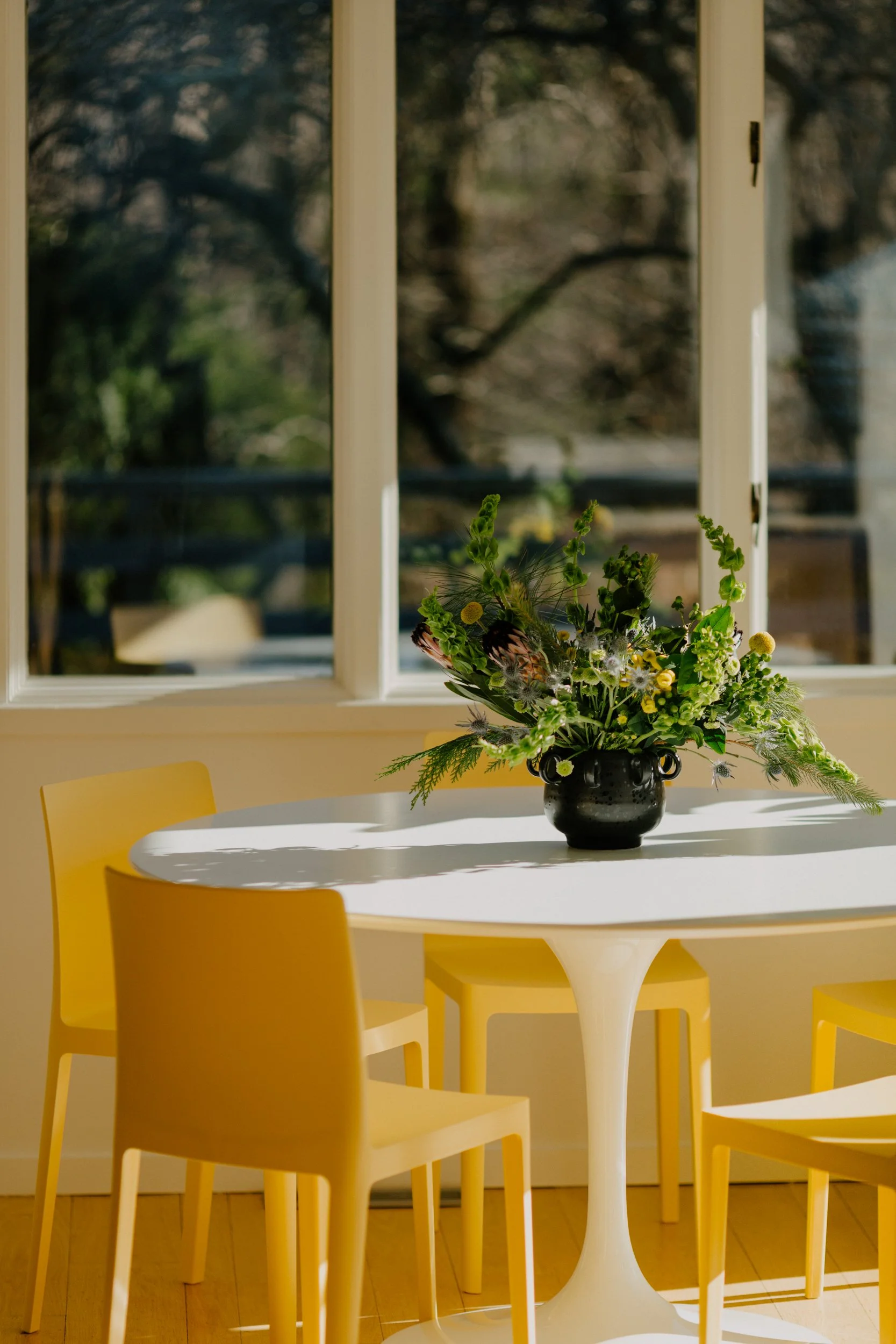 A dining area with a white round table, surrounded by yellow chairs, and a black vase with a green, white, and yellow floral arrangement, in front of a large window letting in sunlight.