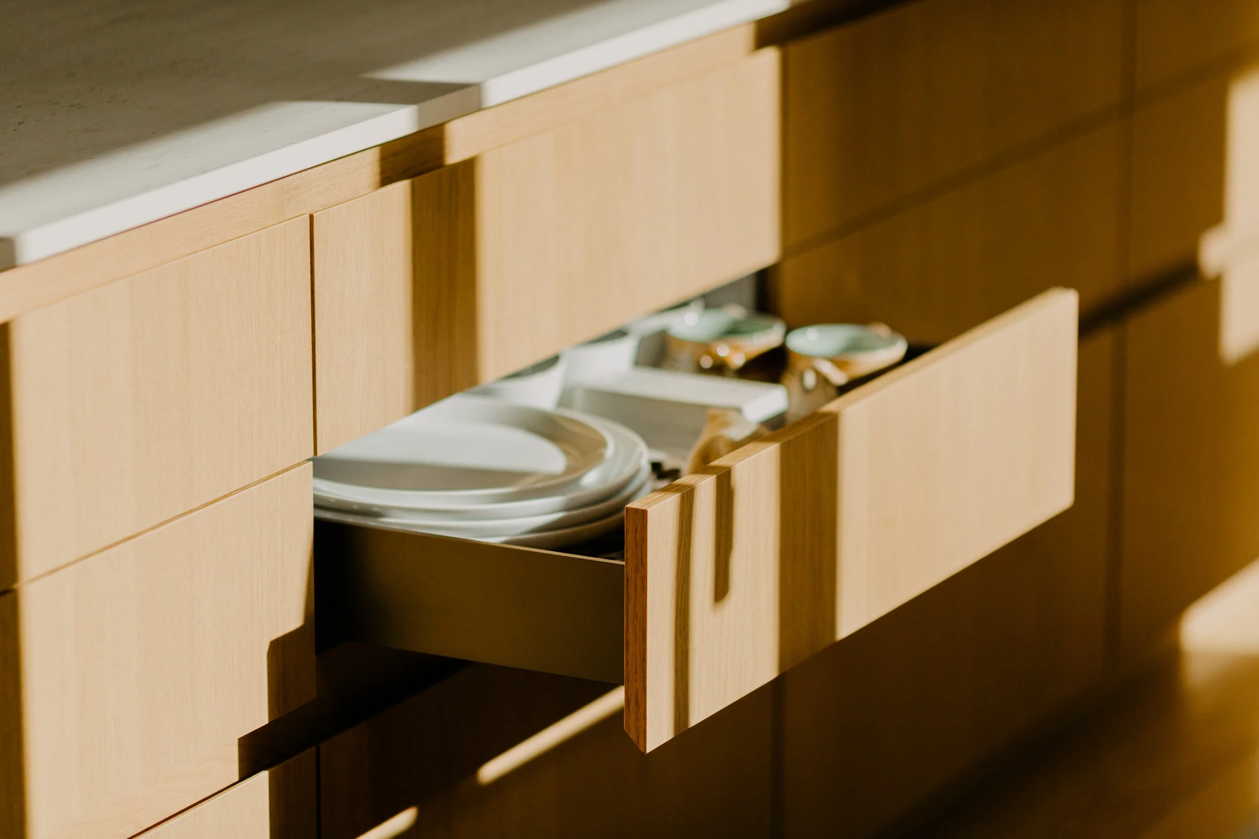 Open kitchen drawer with dishes and bowls inside, sunlight casting shadows.