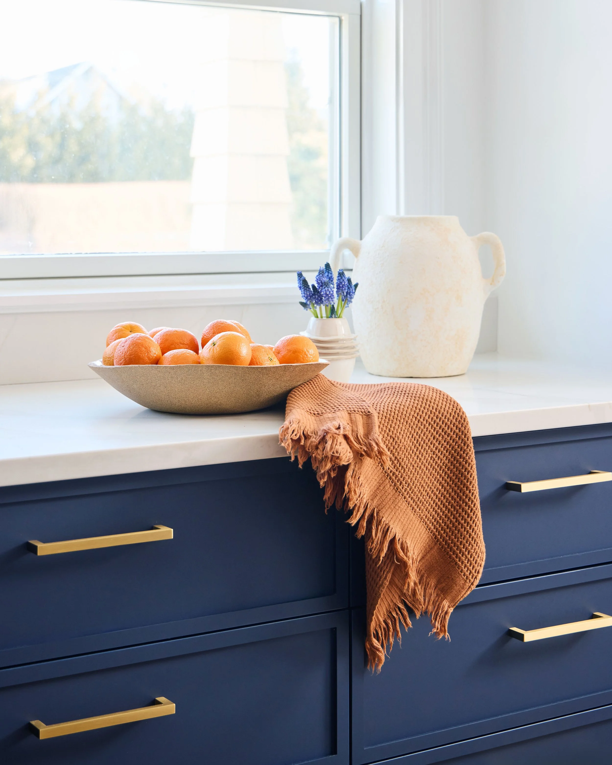 A kitchen counter with a bowl of oranges, a small vase with purple flowers, a white vase, and a brown cloth on dark blue cabinets next to a window.