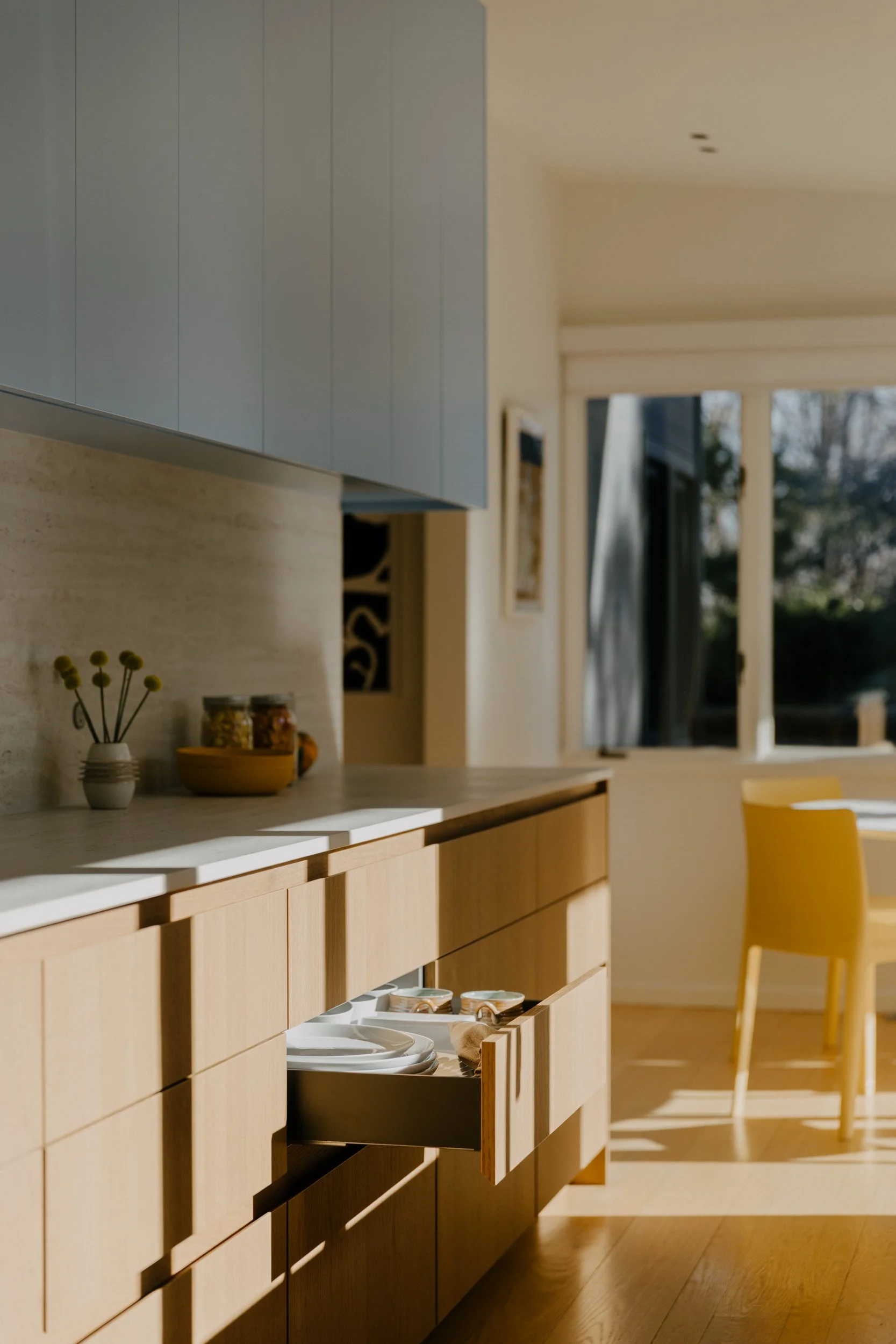A modern kitchen with light wooden cabinets, a countertop with jars and a bowl, and open drawers with plates and bowls inside, illuminated by natural sunlight through a large window.