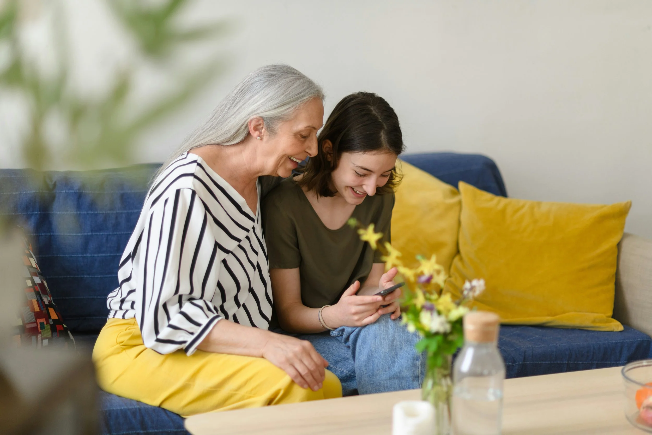 A grandmother and granddaughter sitting on a couch, looking at a smartphone, smiling. The grandmother has gray hair and is wearing a striped shirt, while the girl has dark hair and is wearing a dark green shirt. The room has a white wall, a blue couch with yellow pillows, a wooden table with a vase of yellow flowers, and some candles.