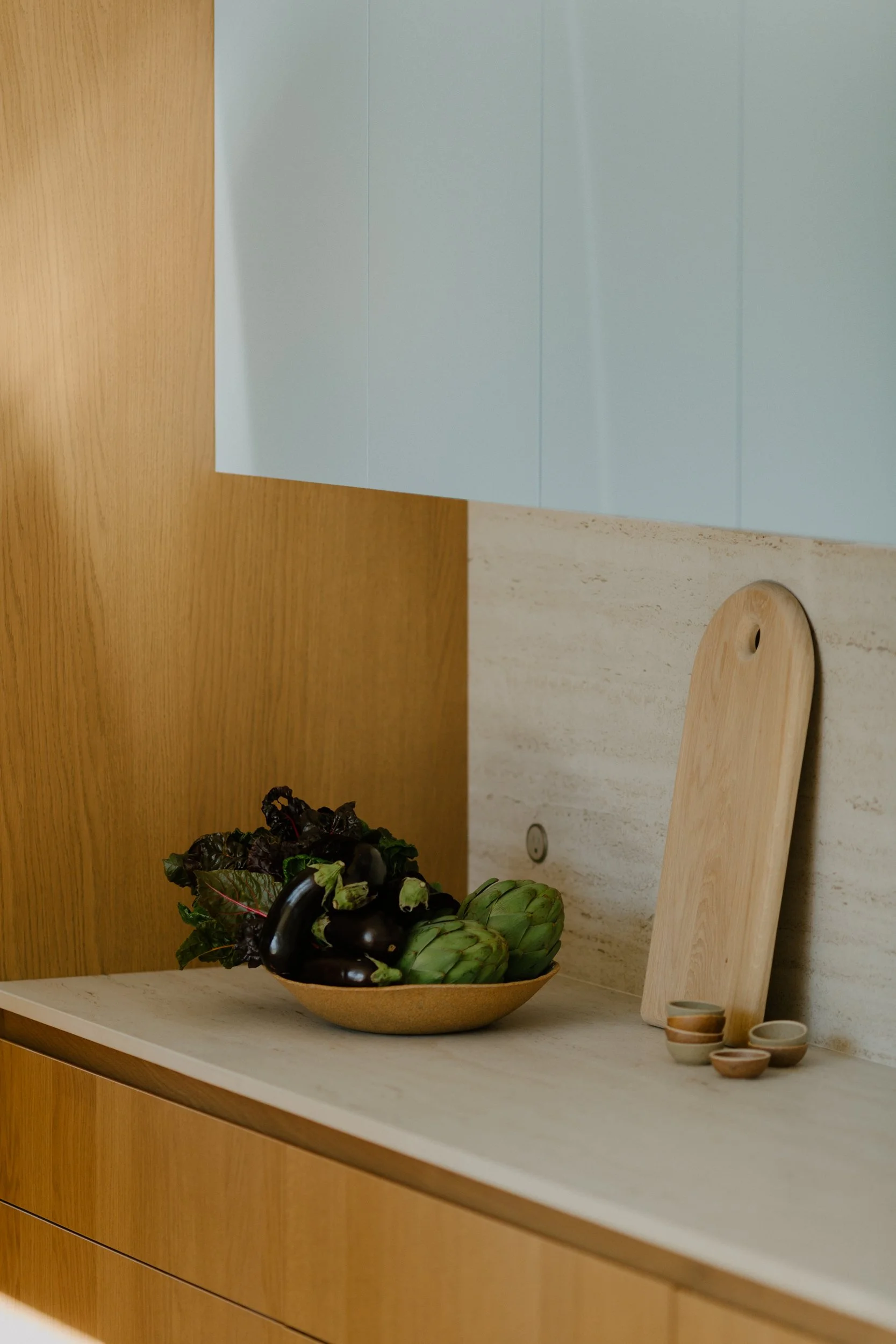 A kitchen countertop with a ceramic bowl of leafy greens and eggplants, a wooden cutting board leaning against the wall, and small bowls nearby. The wall behind is a light-colored stone, with wooden cabinets above and below.