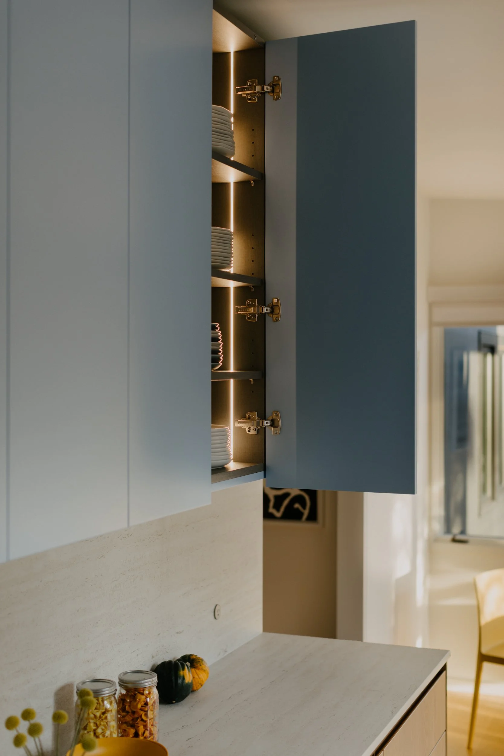 Open blue kitchen cabinet with illuminated interior and stacked dishes inside. Light-colored countertop, small decorative gourds, jars of pasta, and flowers in a vase on it. Part of a dining area with a window and a chair visible in the background.