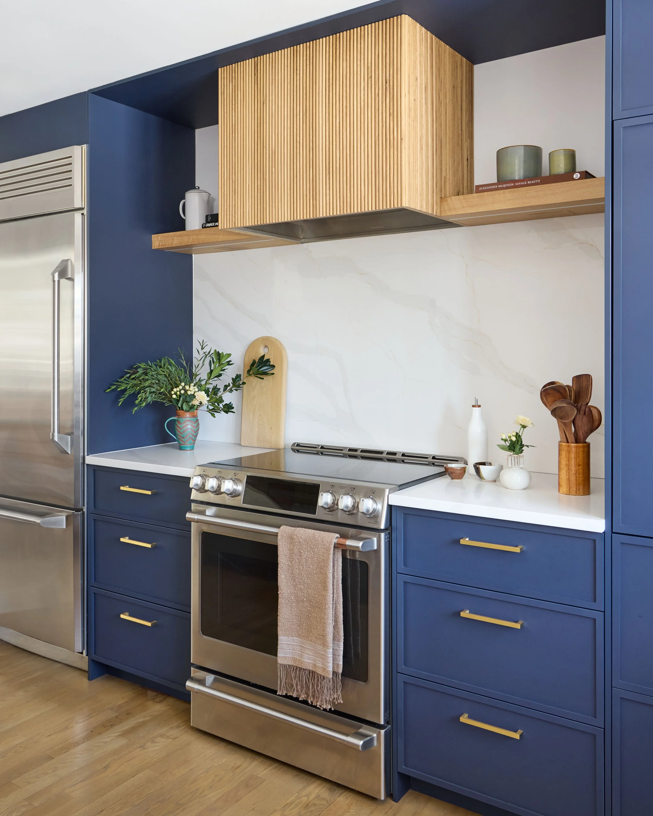 Modern kitchen with blue cabinets, stainless steel oven, white marble backsplash, wooden shelf and decor, including plants and utensils.