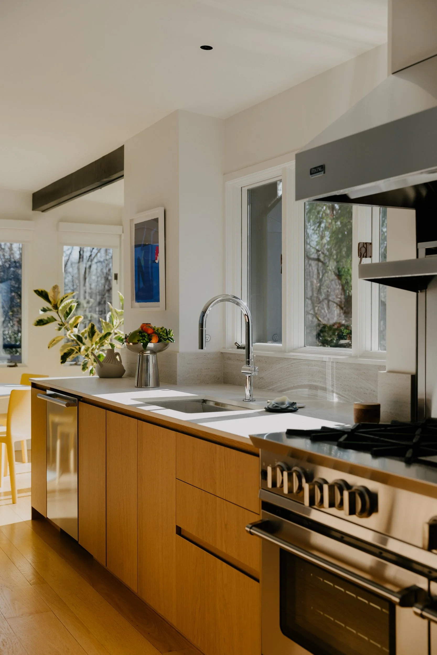 Modern kitchen with white countertops, wooden cabinets, stainless steel appliances, large windows, and a sink with a chrome faucet.