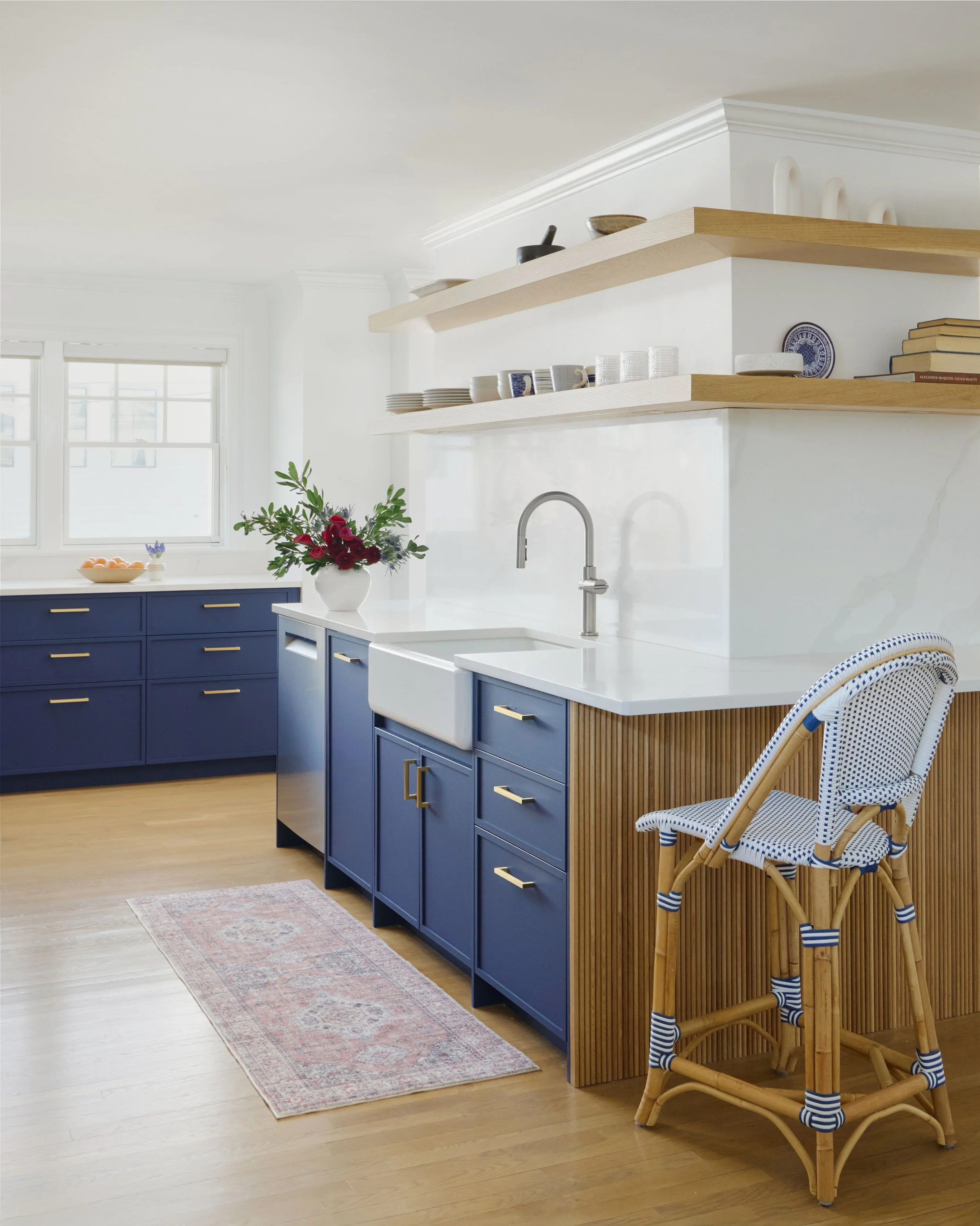 Kitchen with navy blue cabinets, white countertops, open wooden shelves, and a wicker bar stool with a blue and white cushion.
