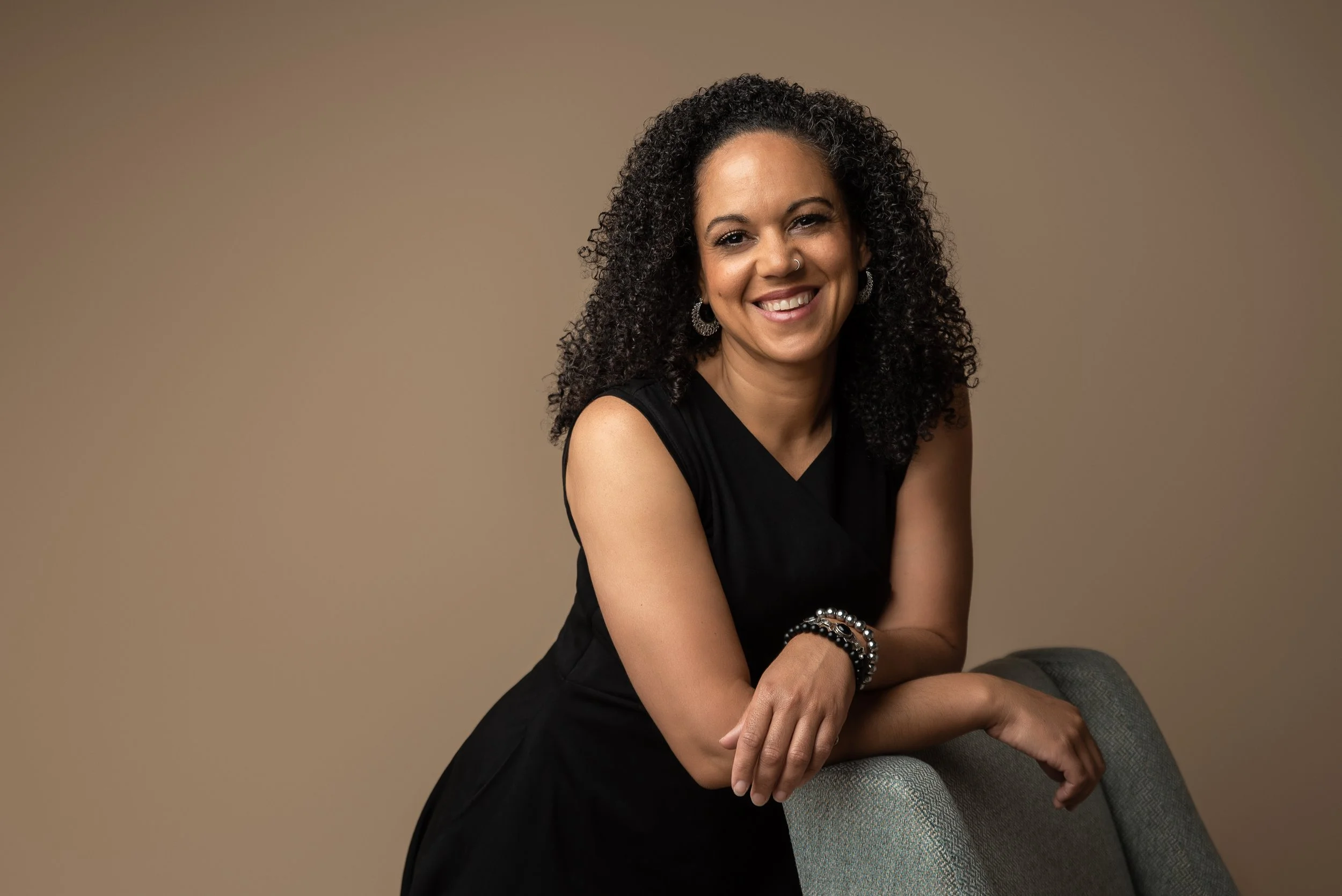 A woman with curly black hair wearing a black sleeveless dress and bracelets, smiling and leaning on the back of a gray chair against a beige background.