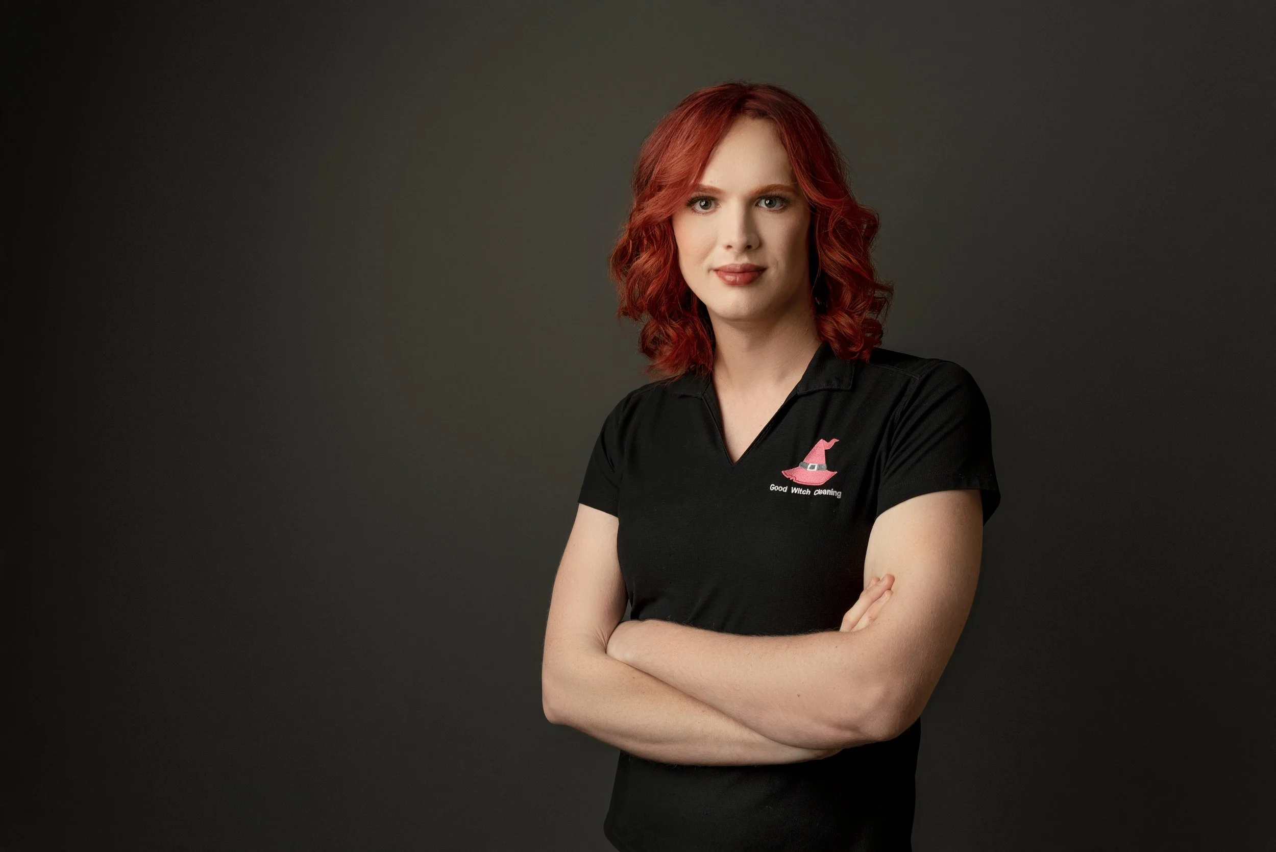 Portrait of a woman with red hair wearing a black polo shirt with a pink witch hat logo and the text 'Good Witch Cleaning' on a dark background.